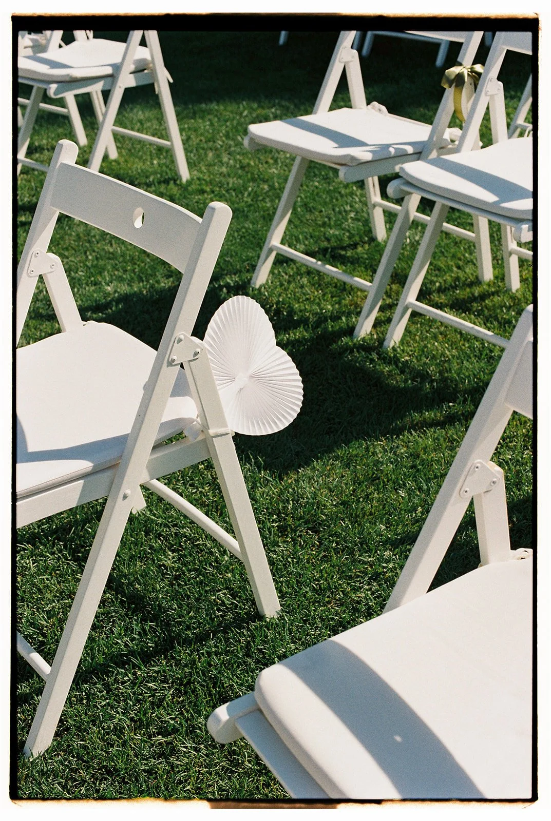 White wedding chairs arranged on a green lawn, decorated with linen fabric and a paper parasol, suggesting an outdoor ceremony, photographed at a wedding in Portugal.