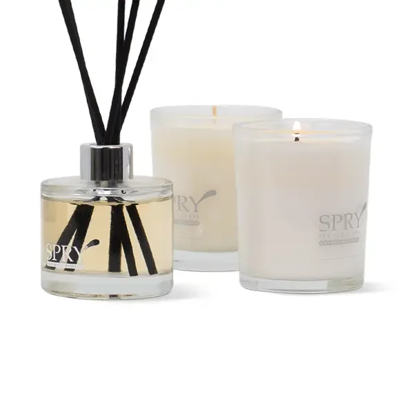 A reed diffuser with black reeds and a glass container, alongside two lit white candles in glass holders, on a white background.