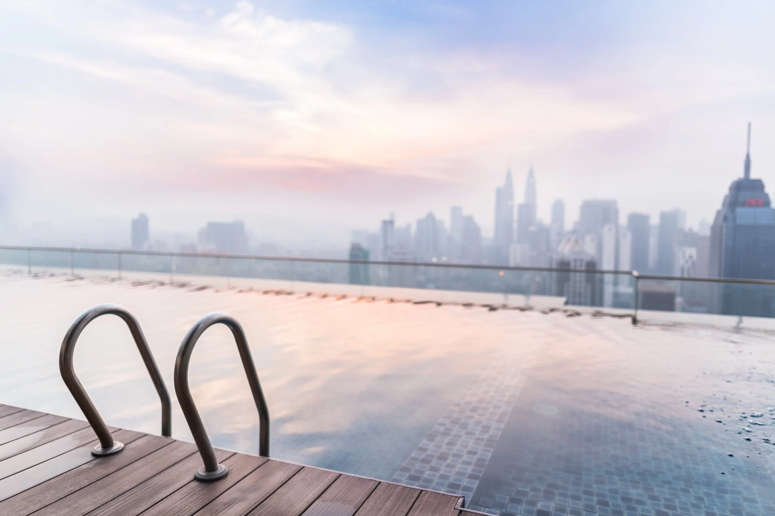 City skyline view from a rooftop pool with ladder and deck in the foreground, hazy sky