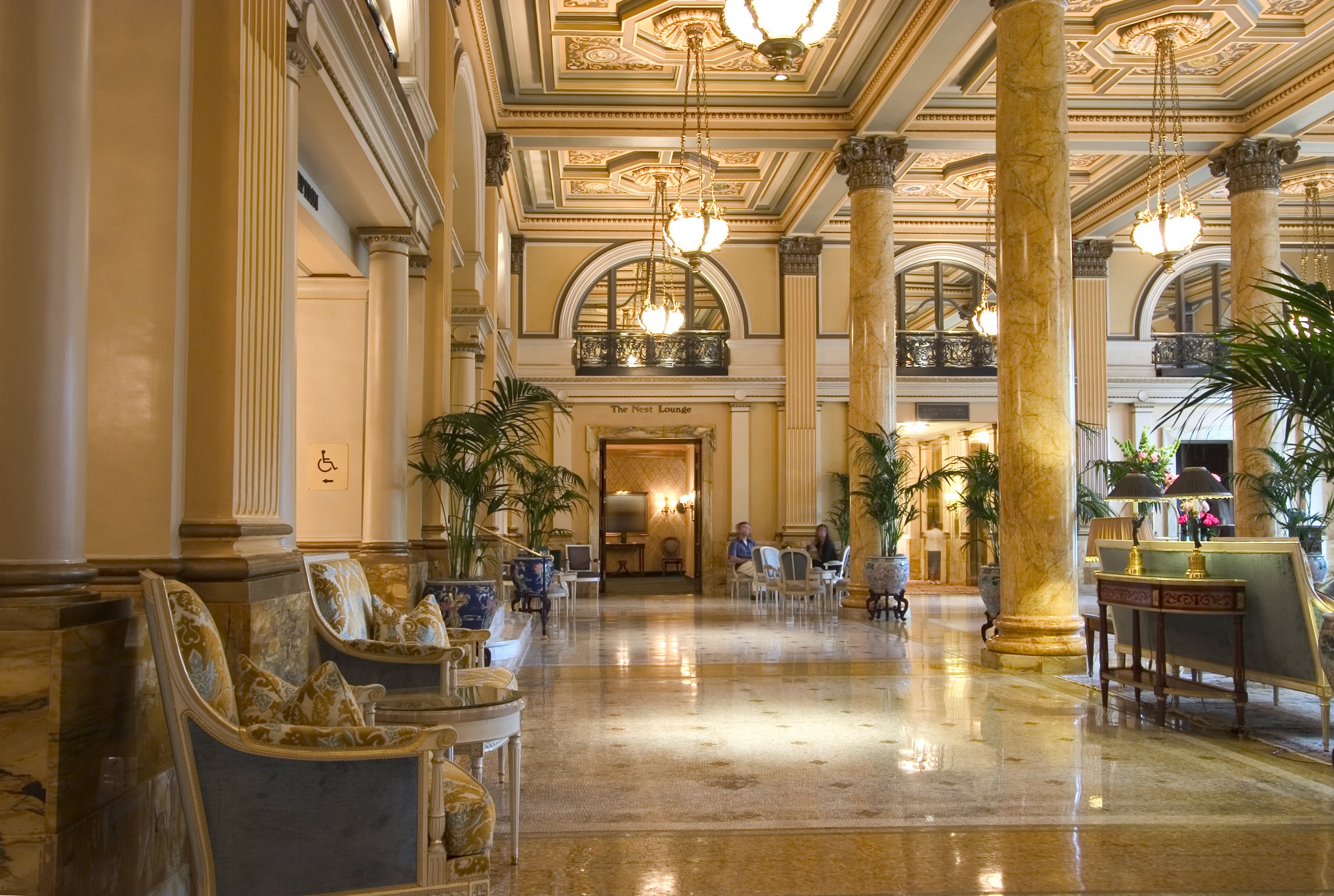 Luxurious hotel lobby with marble floors, tall columns, ornate ceiling with chandeliers, and elegant seating areas.