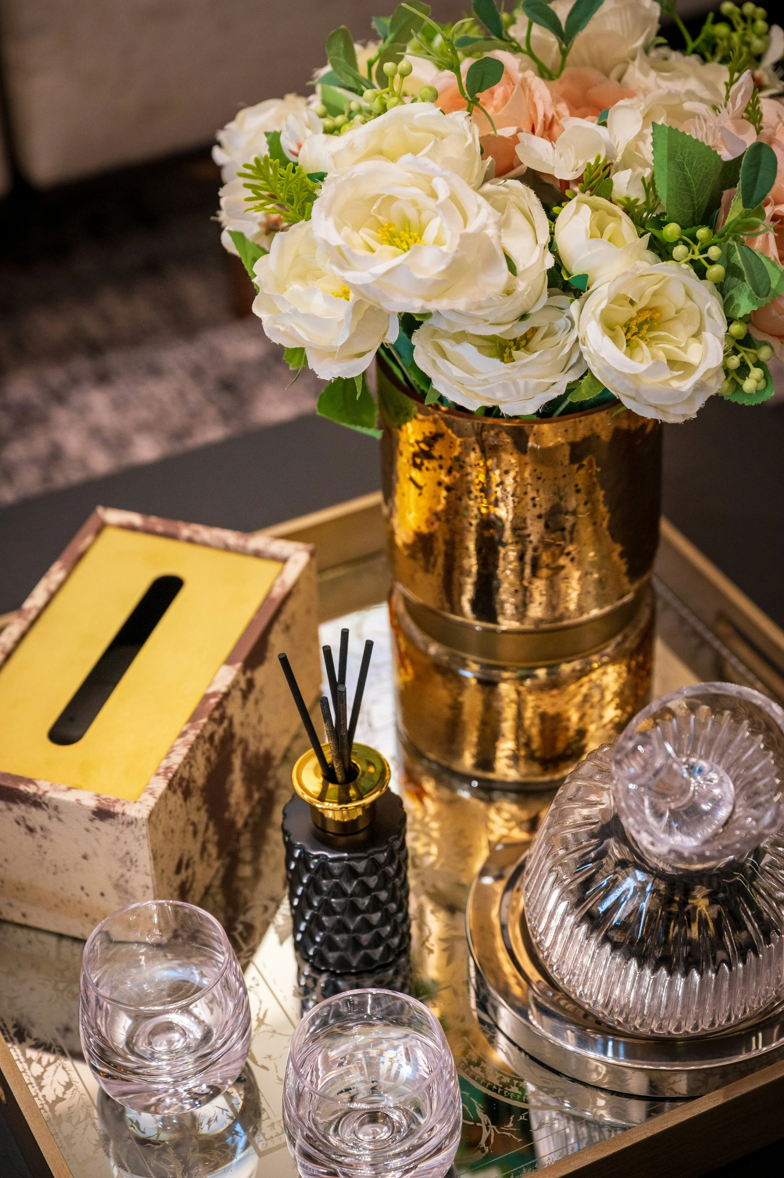 A decorative floral arrangement with white and light pink flowers in a shiny gold vase on a table, surrounded by glasses and decorative items.