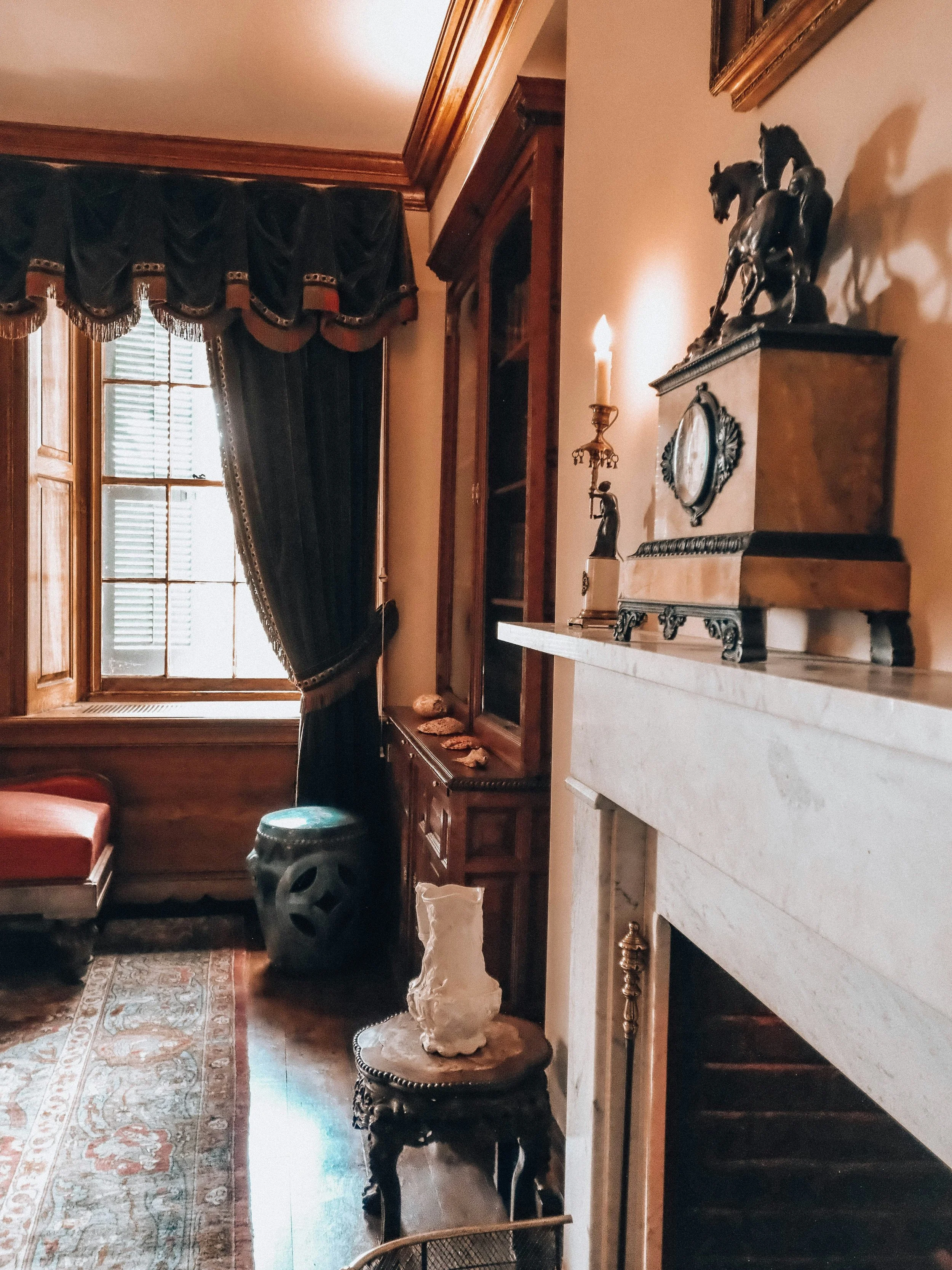 A room with wooden furniture, a window with dark curtains, a carpet with a floral pattern, and decorative items including a horse sculpture and a candle on a holder.