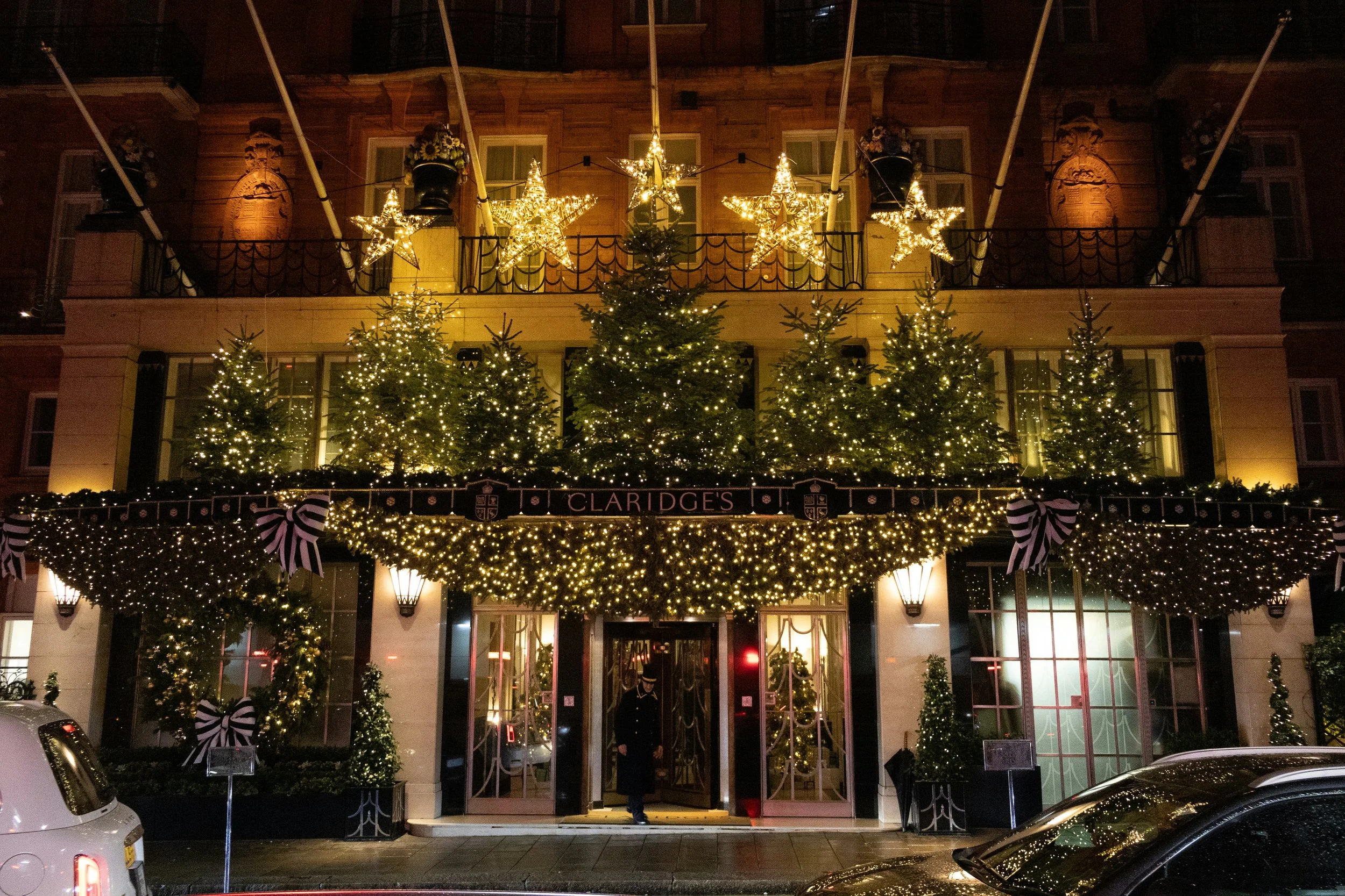 Night view of a decorated building with Christmas lights. The entrance is adorned with Christmas trees with lights, black and white striped bows, and string lights. Five illuminated star decorations are on the balcony railing above the entrance. A person is standing at the doorway.