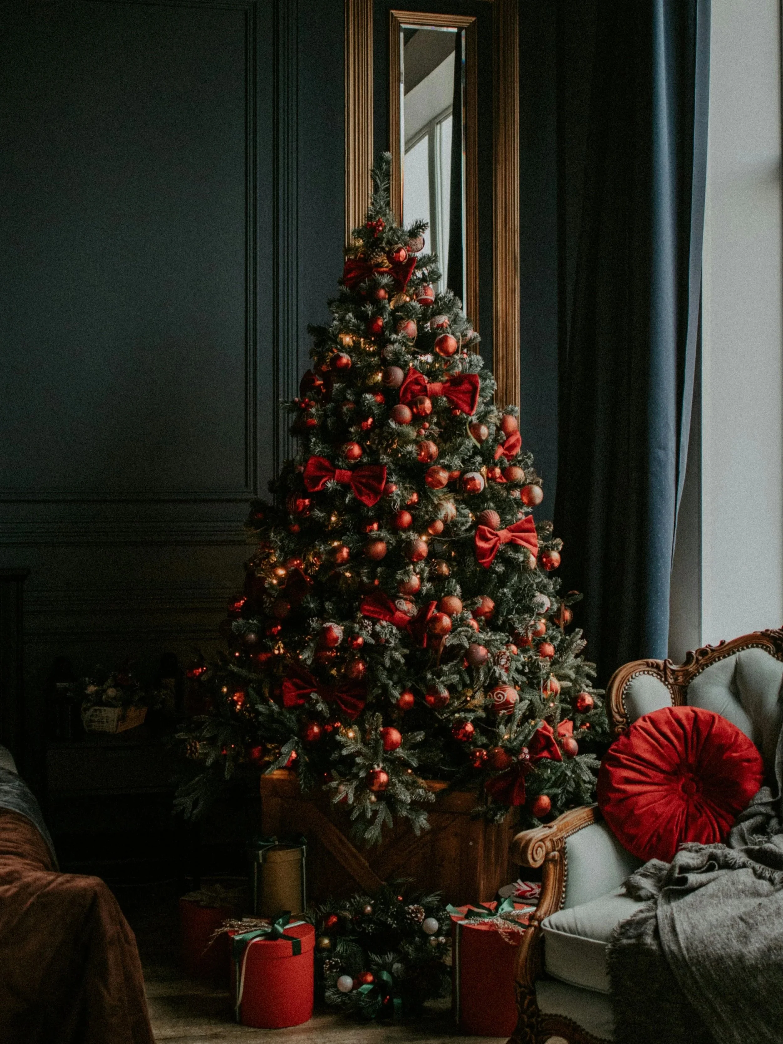 A decorated Christmas tree with red ornaments, bows, and string lights, positioned in a living room against a dark wall and mirror, with wrapped presents at its base and furniture including a vintage sofa and armchair nearby.