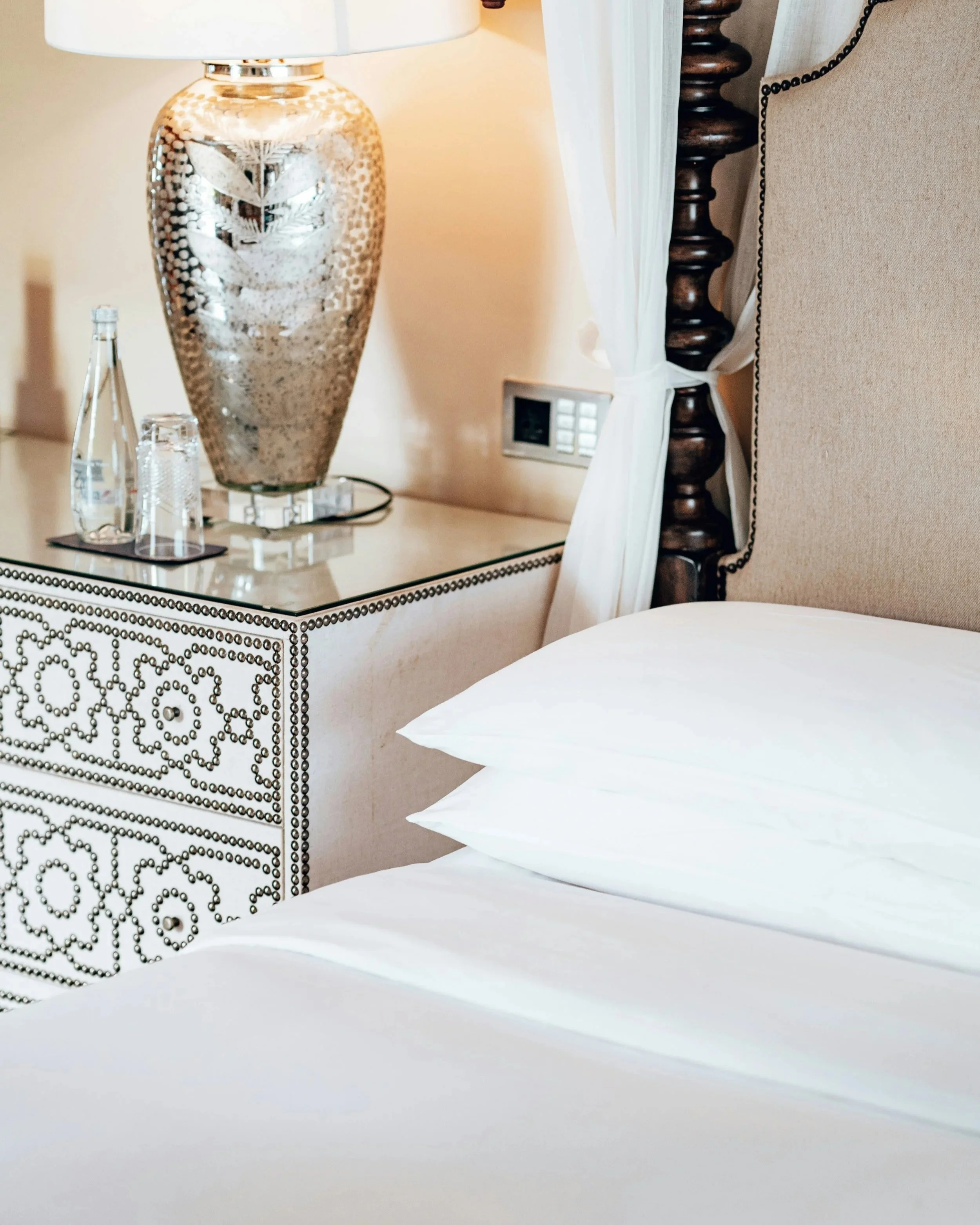 Close-up of a bedside table with a metallic lamp, two glass bottles, and a paper coaster. Part of a bed with white pillows and a beige headboard is visible.