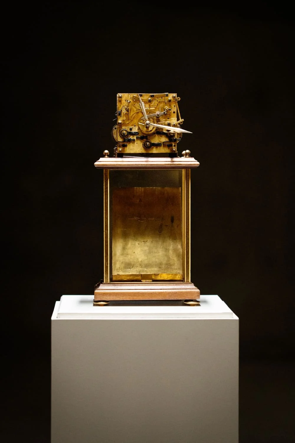 Une horloge ancienne en métal doré exposée sur un socle blanc dans une galerie, avec un fond noir.