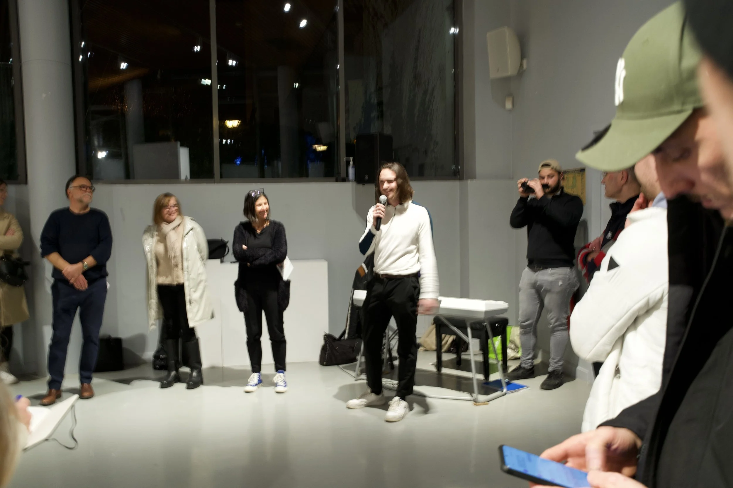 Groupe de personnes lors d'une présentation ou d'un événement social, avec une personne parlant dans un microphone, dans un espace intérieur moderniste.