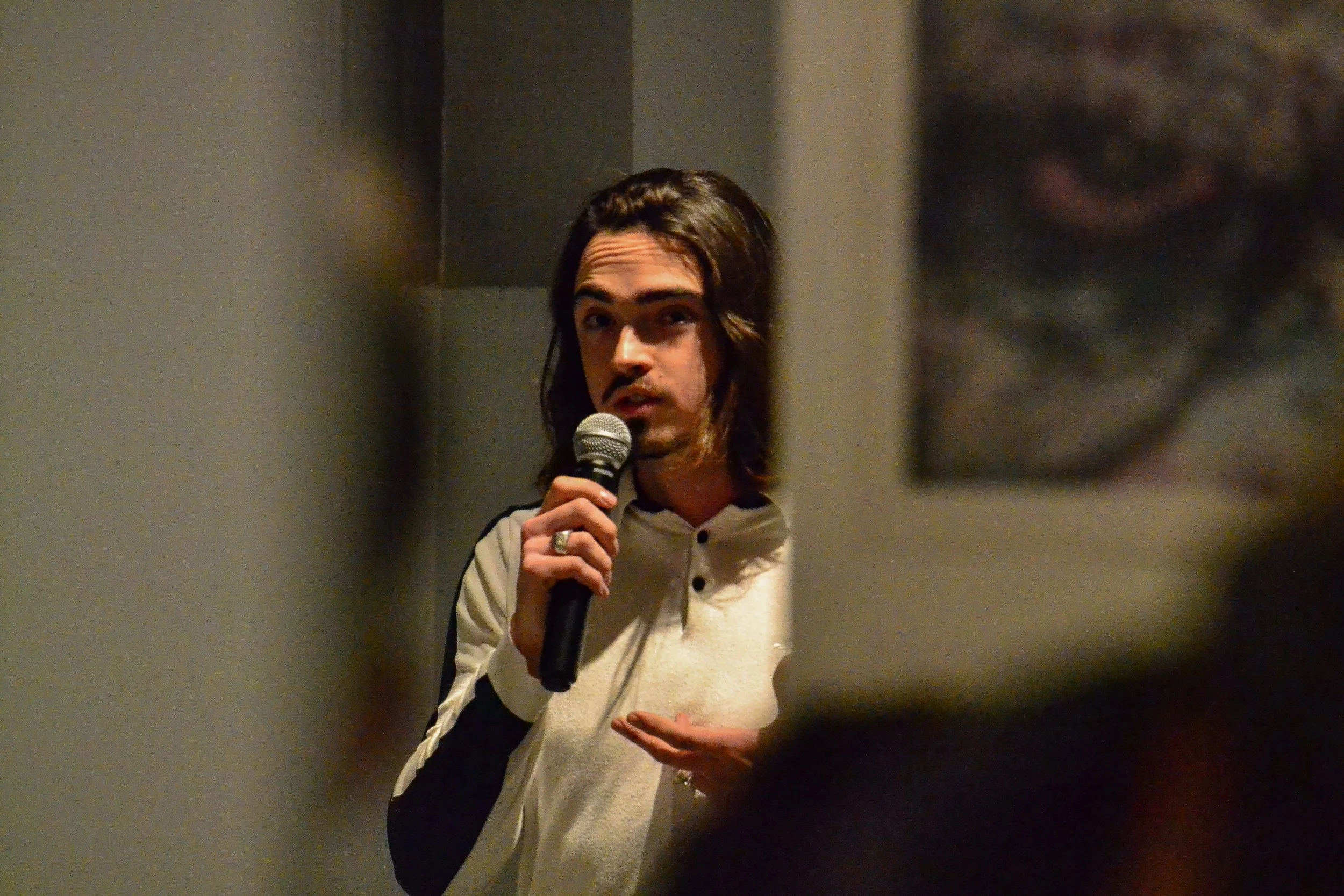 Un homme aux cheveux longs et à la barbe, tenant un microphone, semble parler lors d'une présentation ou d'un discours, dans un espace intérieur avec un fond neutre.