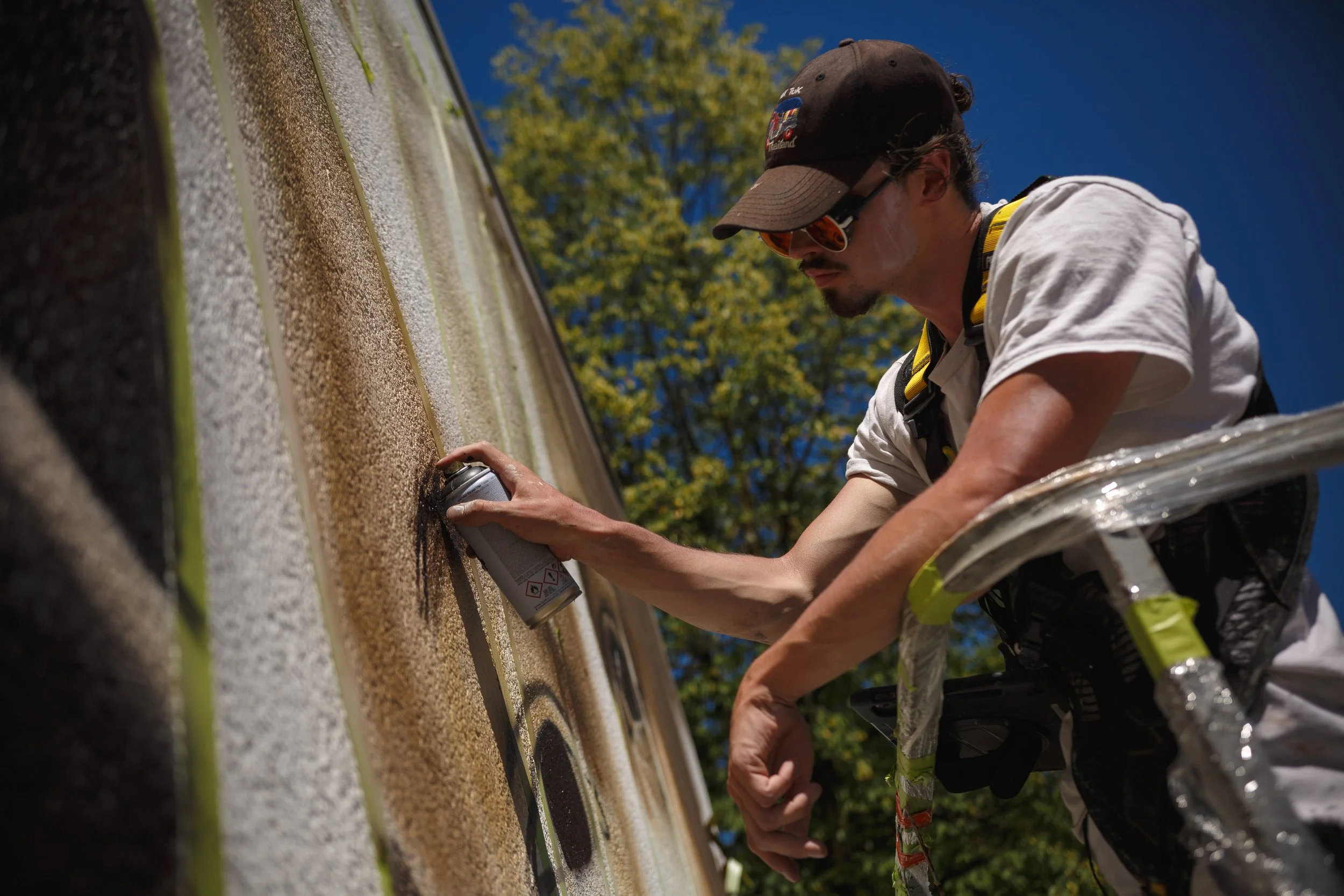 Un homme portant une casquette et des lunettes de soleil, utilisant un spray pour peindre la ligne blanche d'une route en extérieur sous un ciel bleu, avec des arbres en arrière-plan.