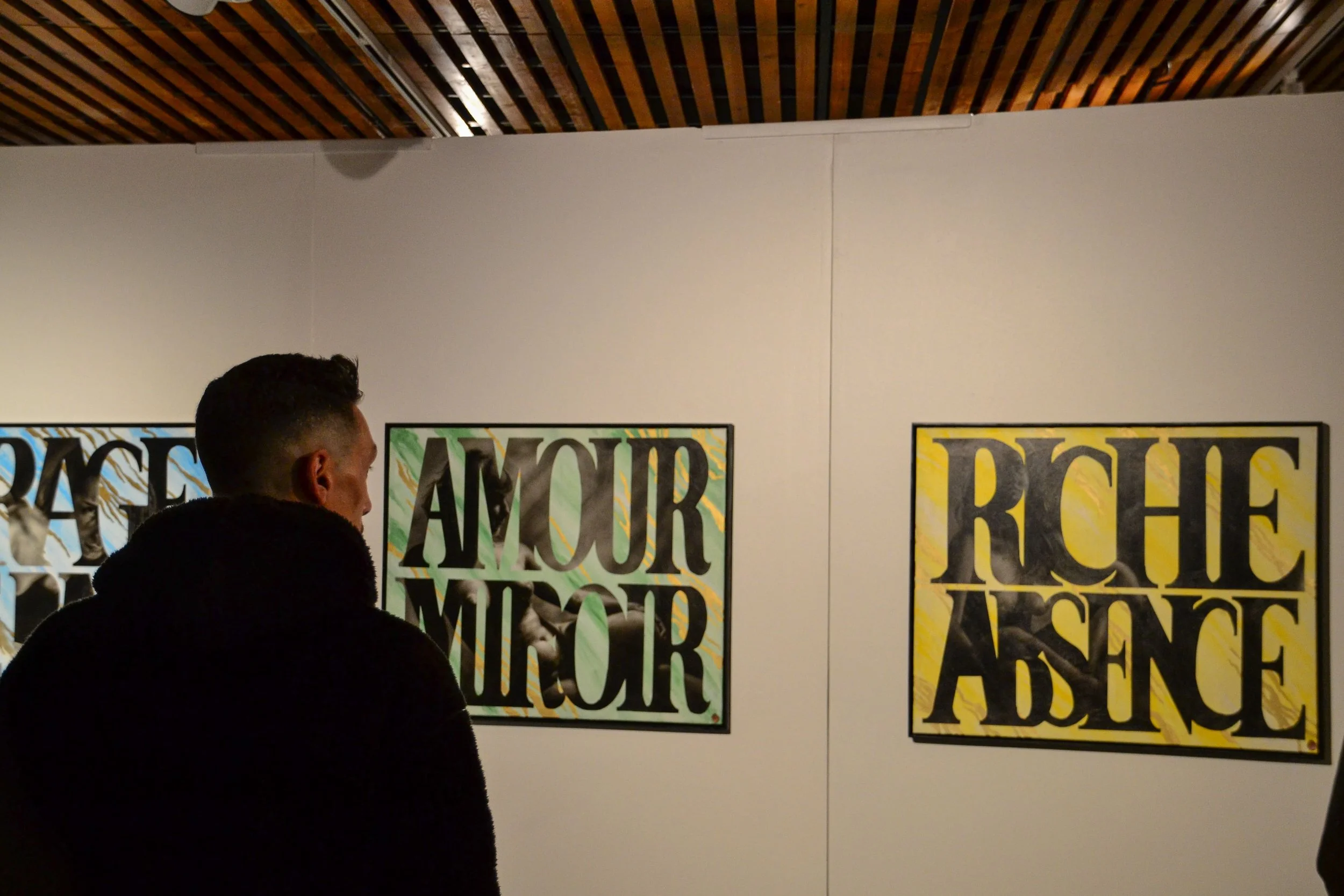 Exposition d'art avec plusieurs affiches colorées et un mur blanc, un homme vu de dos observe l'œuvre, plafonnier en bois visible au plafond.