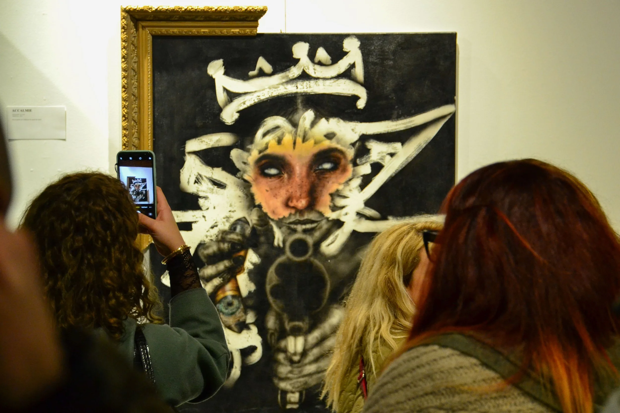 Une œuvre d'art représentant une figure humaine avec un visage déformé, portant un chapeau orné de motifs, exposée dans un musée avec plusieurs personnes qui prennent des photos ou regardent l'œuvre.