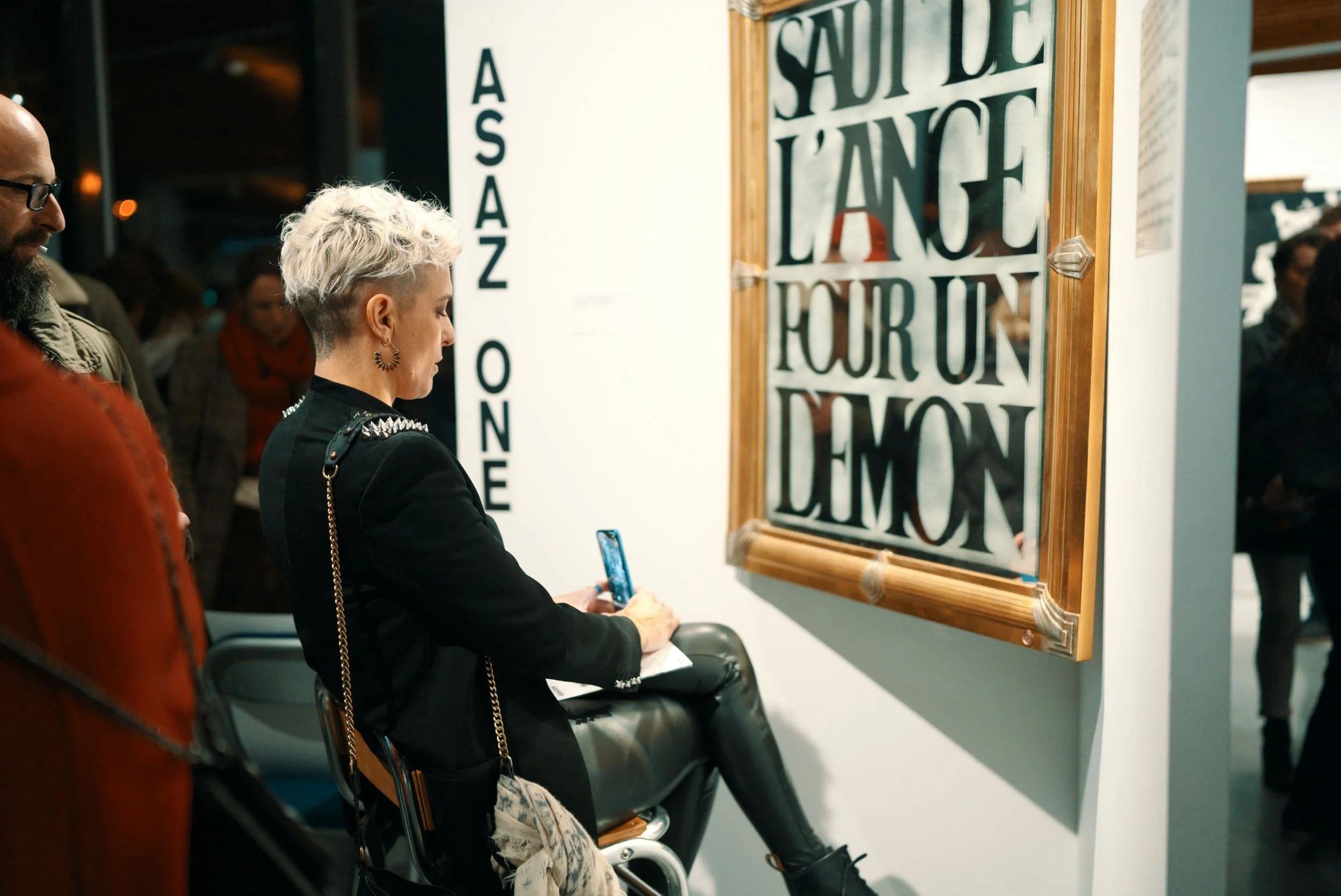 Une femme avec des cheveux courts, blancs et bouclés, assise dans une galerie d'art, prenant une photo avec son téléphone, à côté d'une œuvre d'art accrochée au mur.