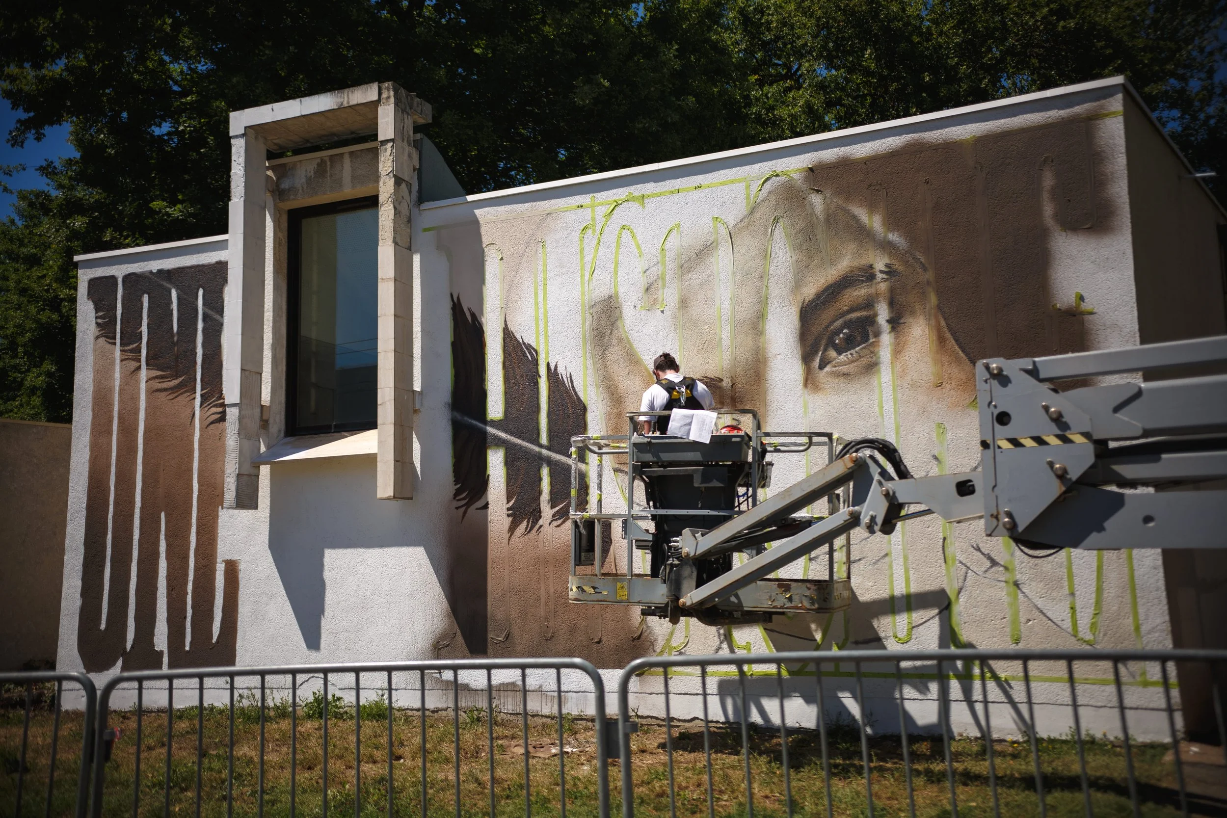 Un artiste peint un grand mural réaliste d'un visage humain sur un mur blanc, utilisant une plateforme élévatrice, avec un ciel clair et des arbres en arrière-plan.