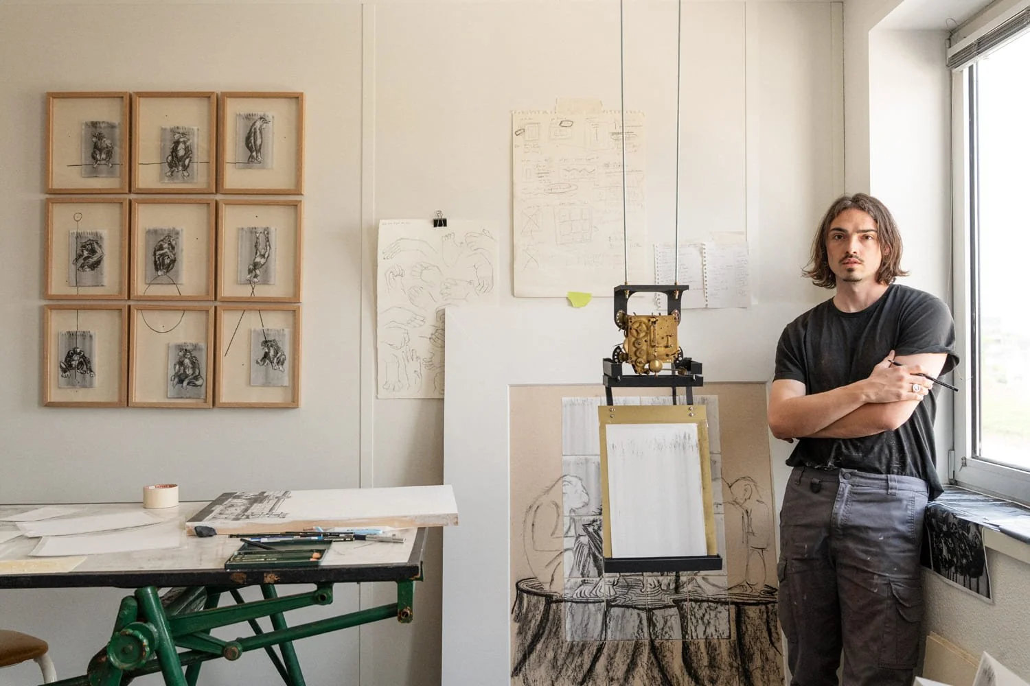 Un homme debout dans un atelier d'artiste, près d'une fenêtre, avec des dessins et des œuvres d'art accrochés au mur et à proximité, un tableau blanc, des croquis, une table avec des outils, et un support pour les œuvres.