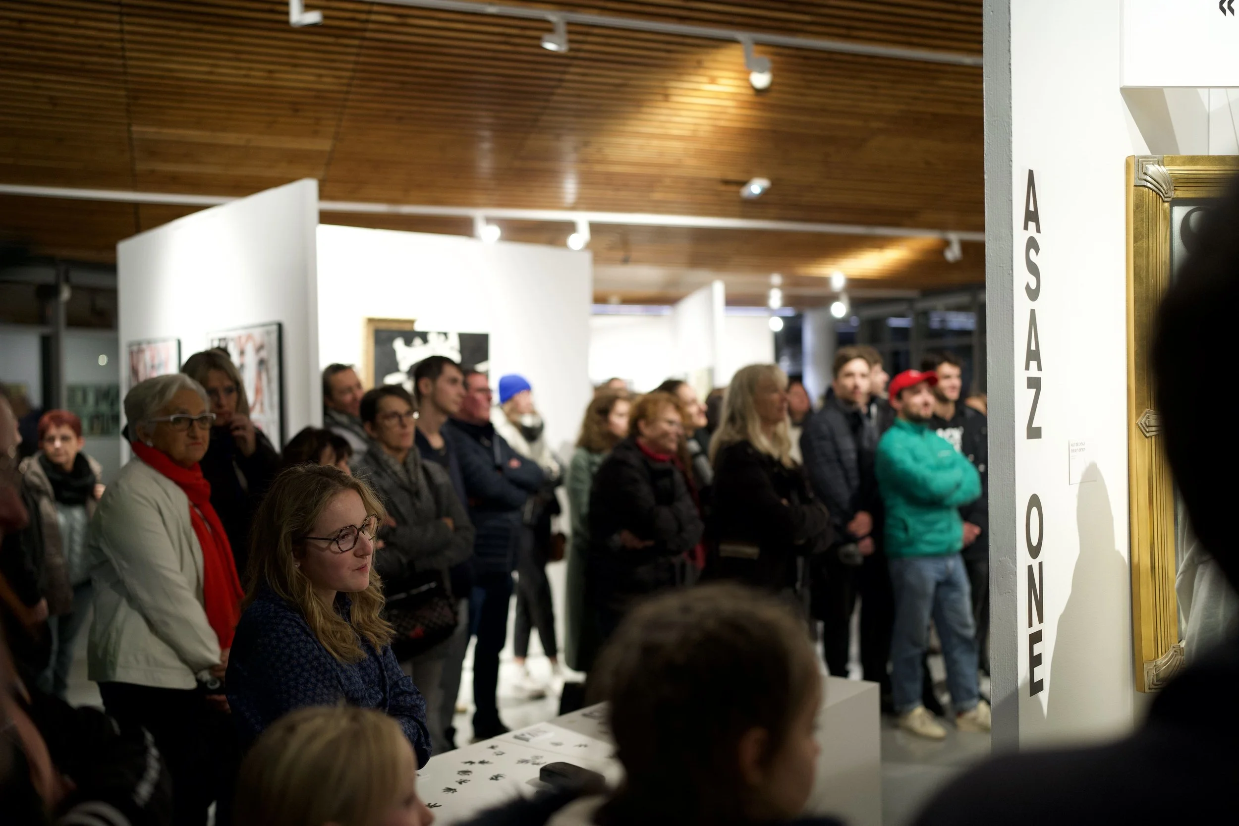 Public réunie dans une galerie d'art, regardant une exposition de portraits et d'œuvres d'art sur les murs.