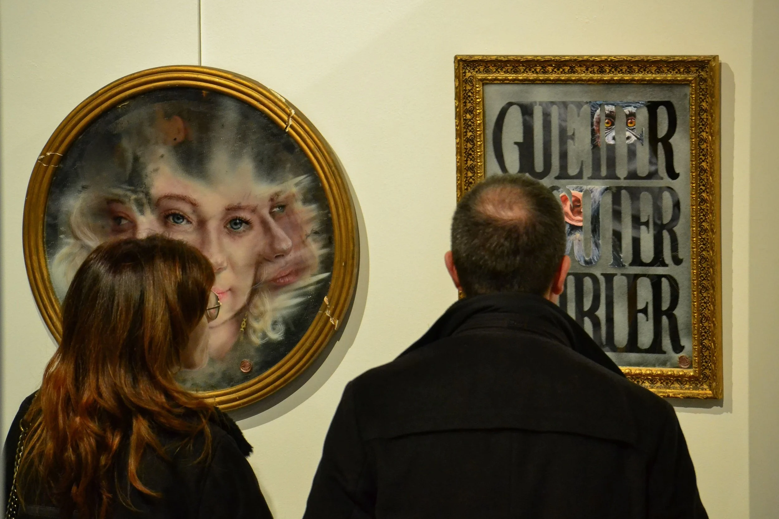 Trois personnes regardent deux œuvres d'art dans une galerie, un portrait d'une femme avec des cheveux blonds et un panneau avec une photo de lynx et les mots 'Guerre' et 'Ours'.