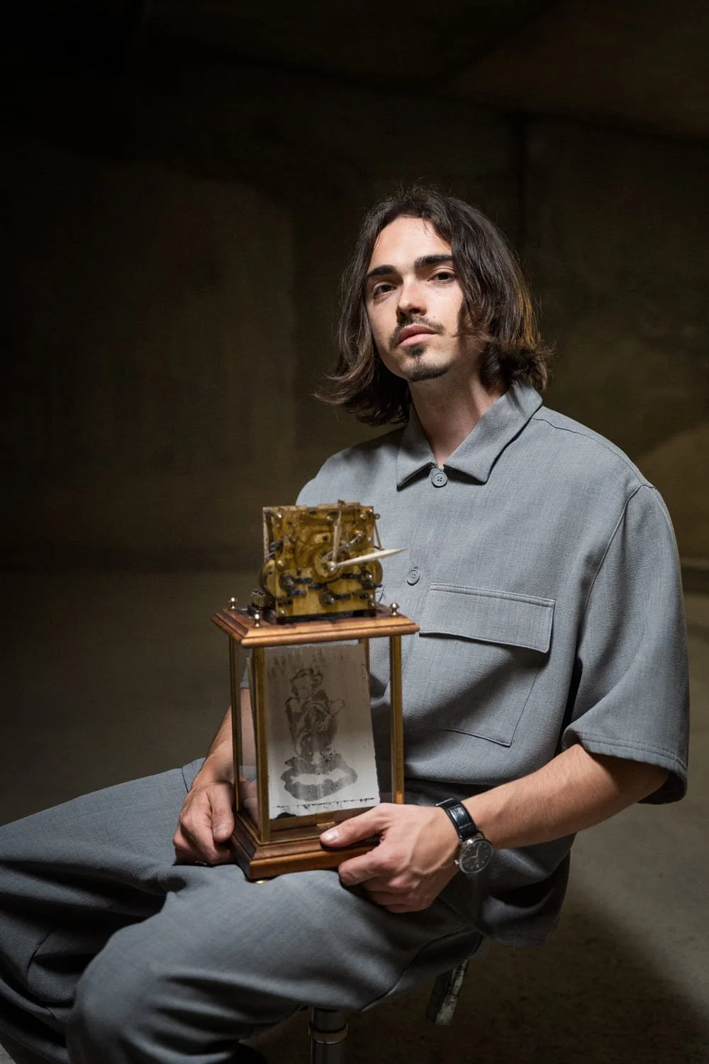 Un homme avec des cheveux longs et une barbe, portant un pull gris, tient une montre à son poignet gauche, et est assis tenant une horloge ancienne avec un cadre en bois et une pièce mécanique à l'intérieur, dans un lieu sombre.