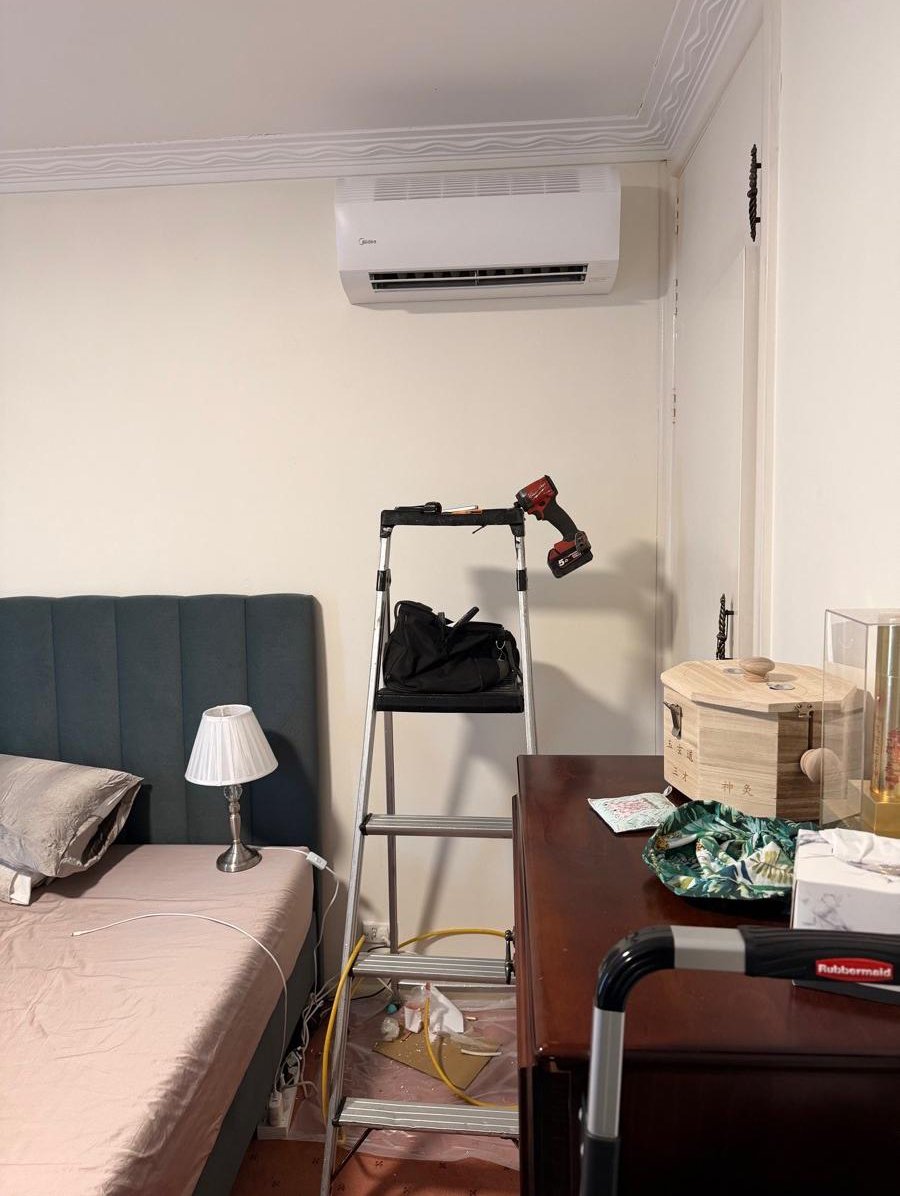 A bedroom with a bed, a small table lamp, a ladder with a power drill, and a dresser with various items on it, including a wooden box, a patterned cloth, and boxes.