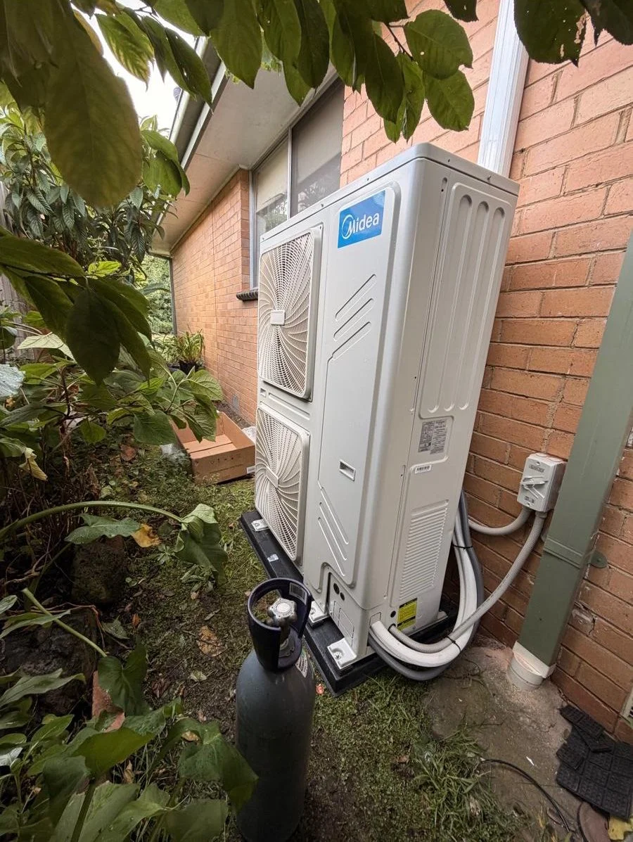 An outdoor air conditioning unit installed on a concrete platform next to a brick house wall, with greenery and plants nearby.