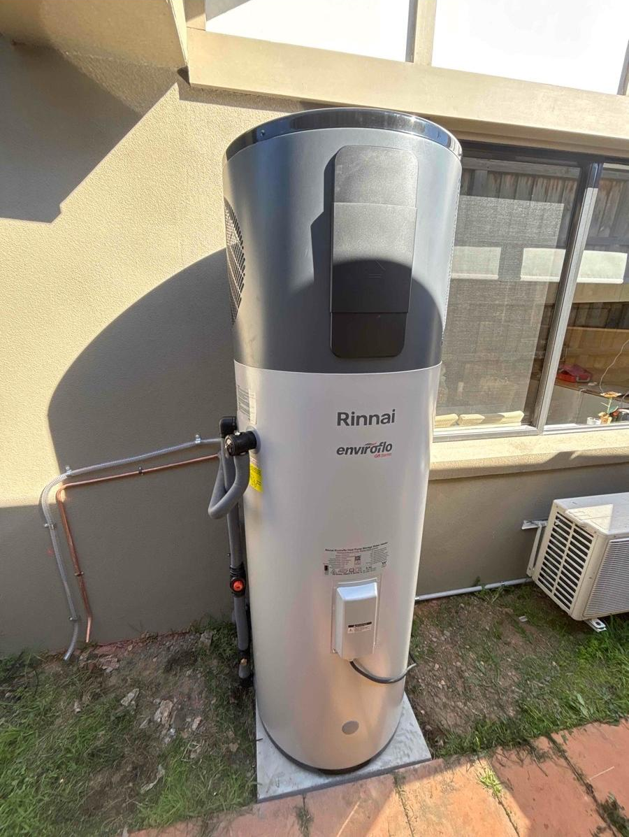 An outdoor Rinnai Enviroflo tankless water heater installed next to a house wall, with pipes connected on the side and an air conditioning unit nearby.