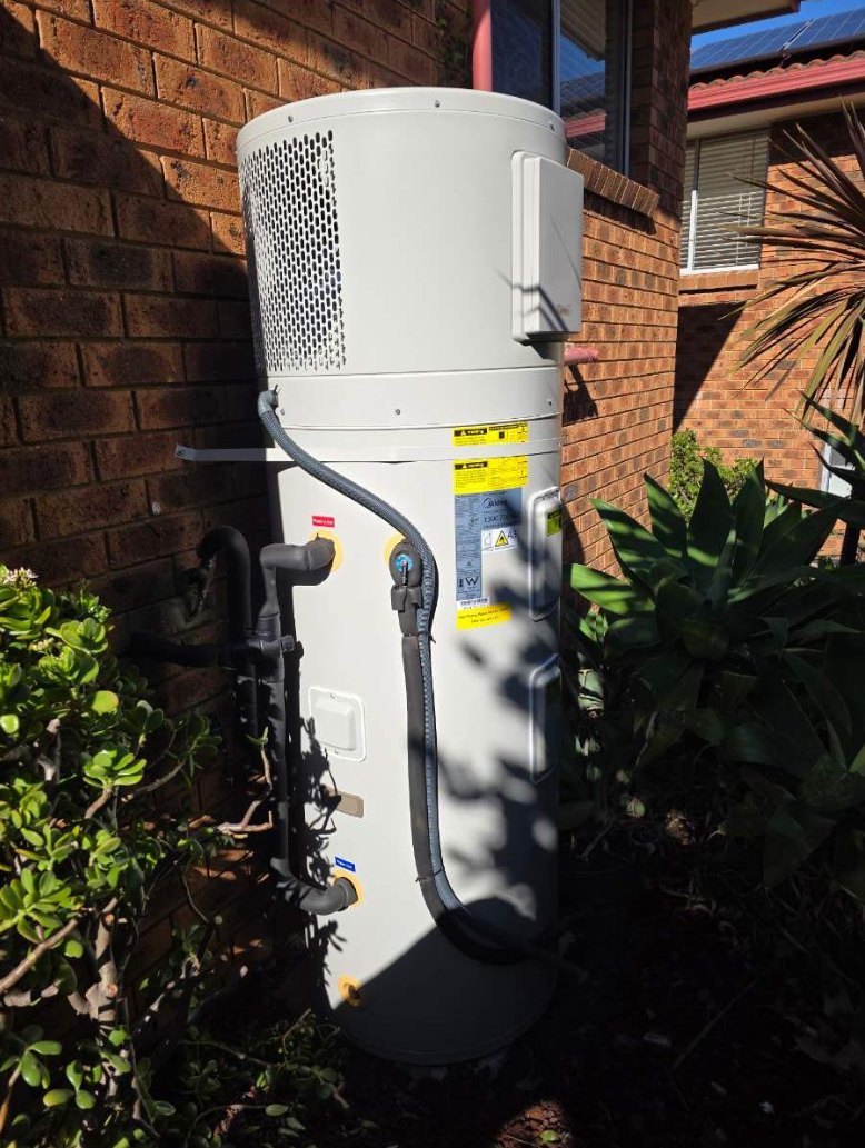 White outdoor water heater tank installed next to a brick wall with greenery and plants around.