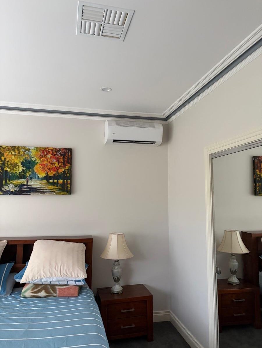 Bedroom corner with a bed, bedside table, lamp, wall art, air conditioner, mirror, and ceiling vents.