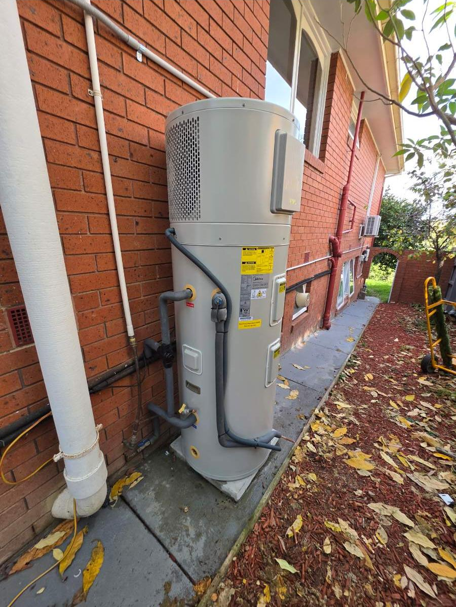 An outdoor water heater installed alongside a brick wall of a house with pipes and vents, and fallen leaves on the ground.