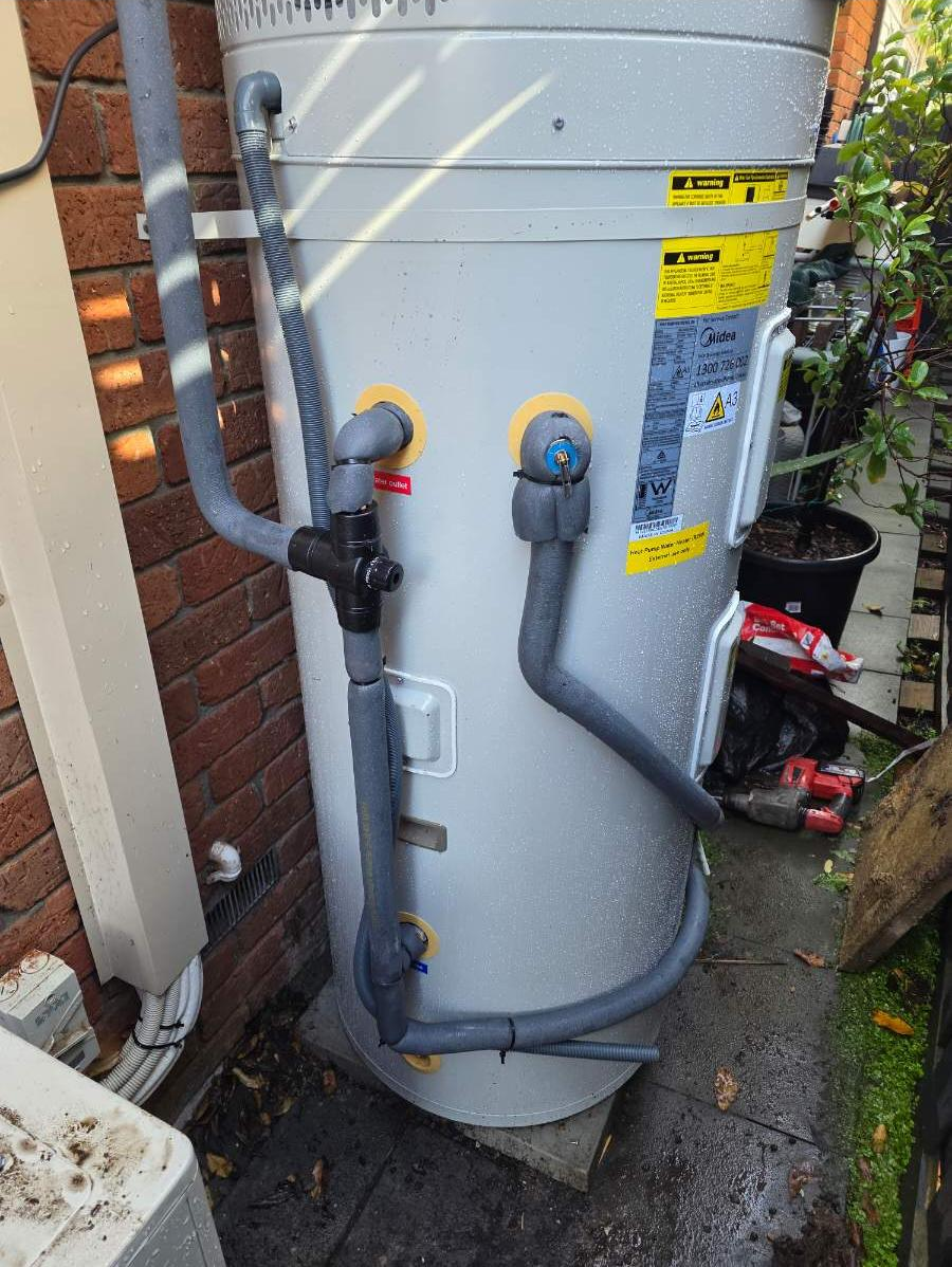 An outdoor hot water heater with pipes and hoses connected, situated next to a brick wall, with tools and a potted plant nearby.