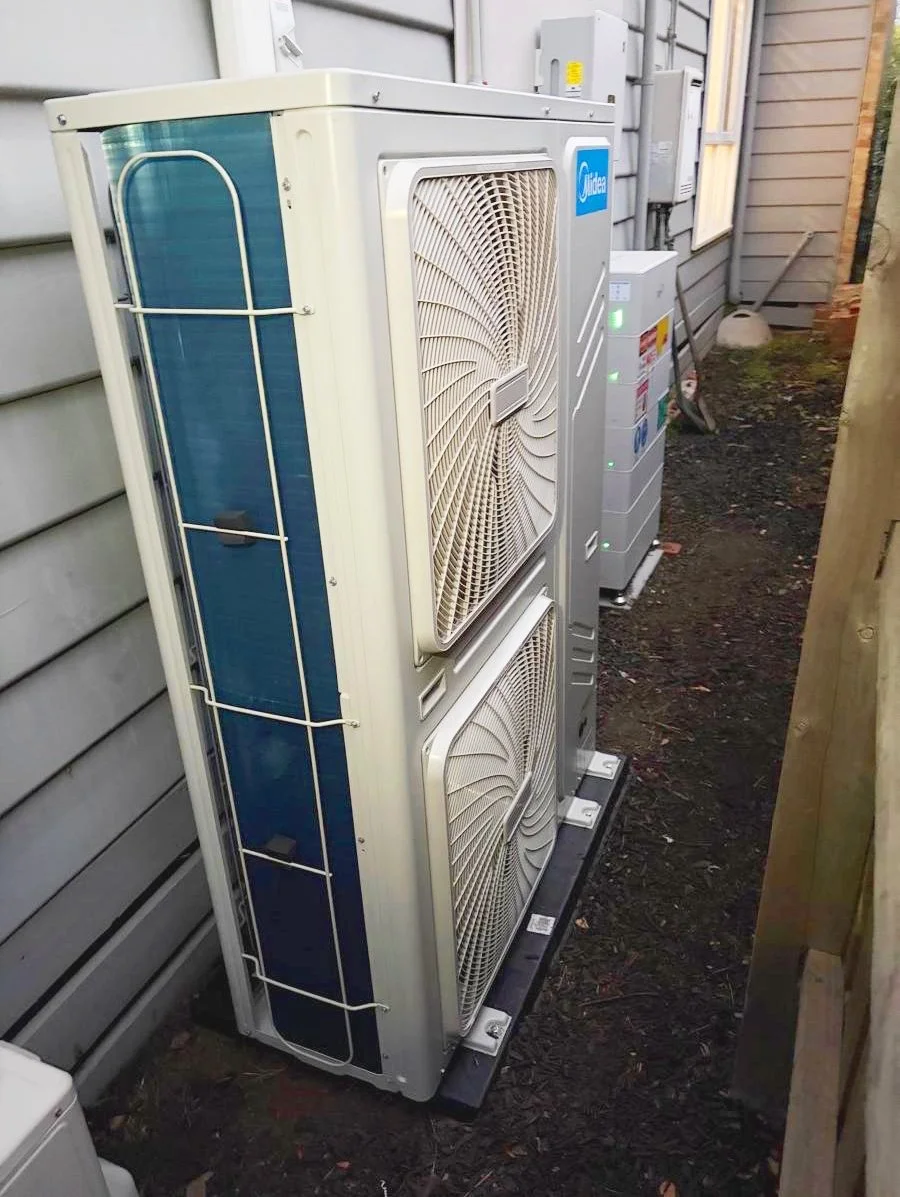 Outdoor air conditioning unit with two large fans and a blue logo, installed next to a house wall.