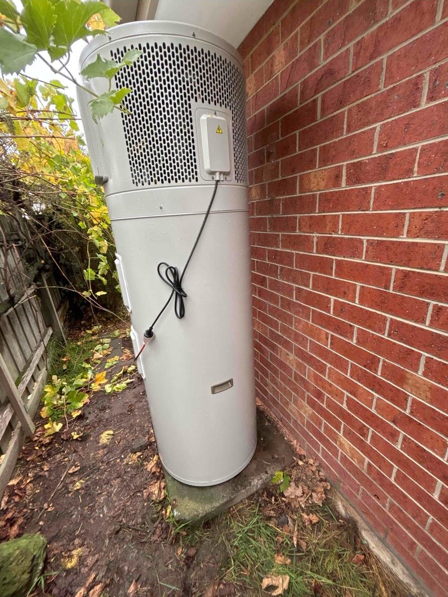 An outdoor HVAC unit or air conditioning compressor next to a brick wall with some greenery and leaves on the ground nearby.