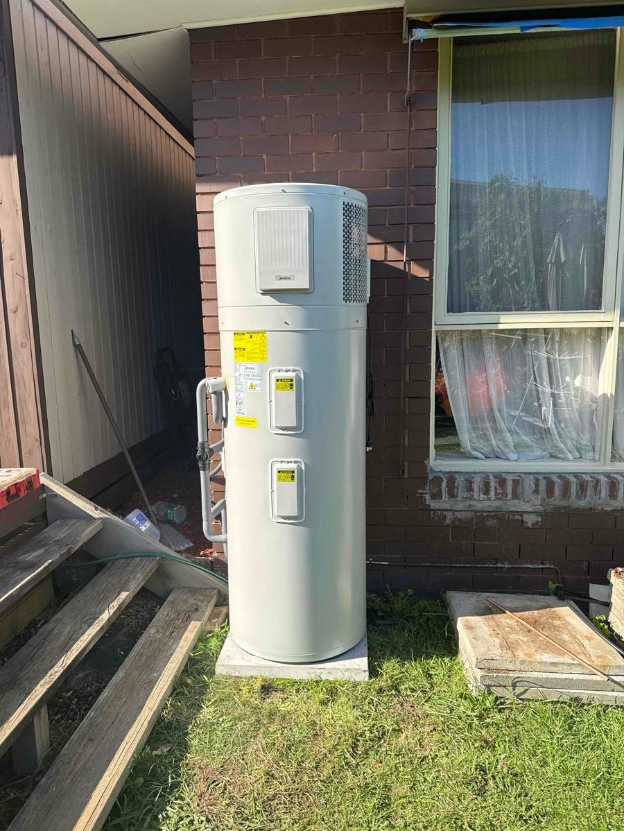 An outdoor Rennai water heater tank installed next to a brick house with an adjacent window with curtains. The tank is on a concrete pad in a grassy area, with some construction materials and tools nearby.