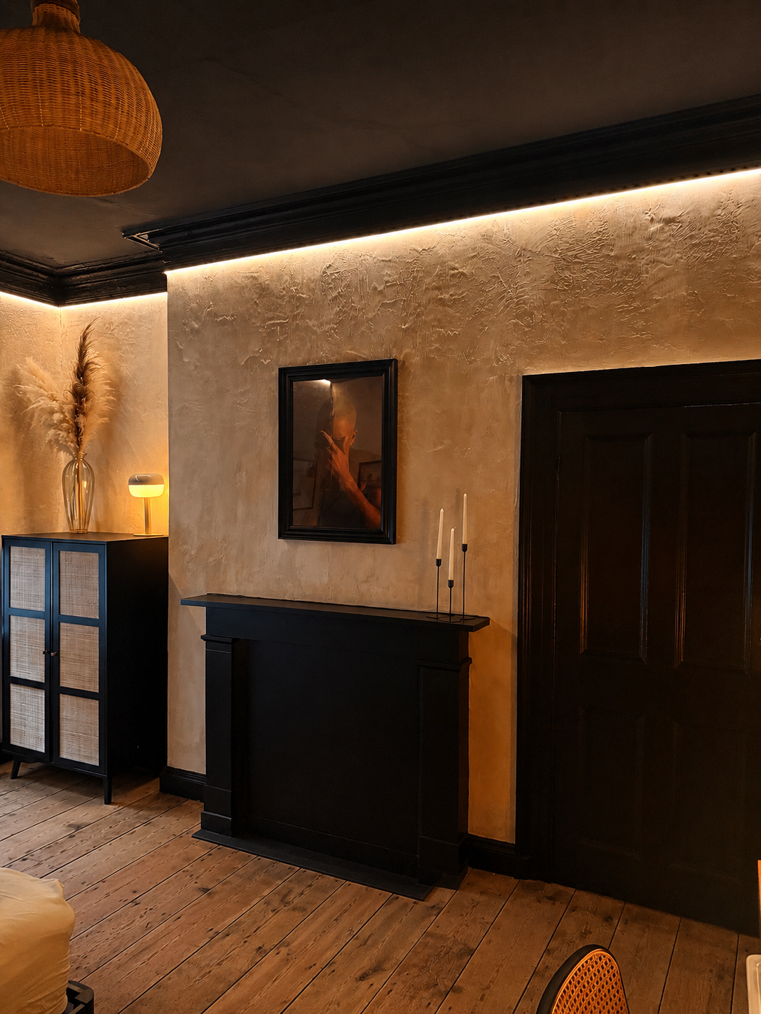 Living room corner with beige textured wall, black fireplace mantel, black door, black cabinet with woven detail, vase with pampas grass, small lamp, and wicks in candlesticks on the mantel, illuminated by warm layered lighting.