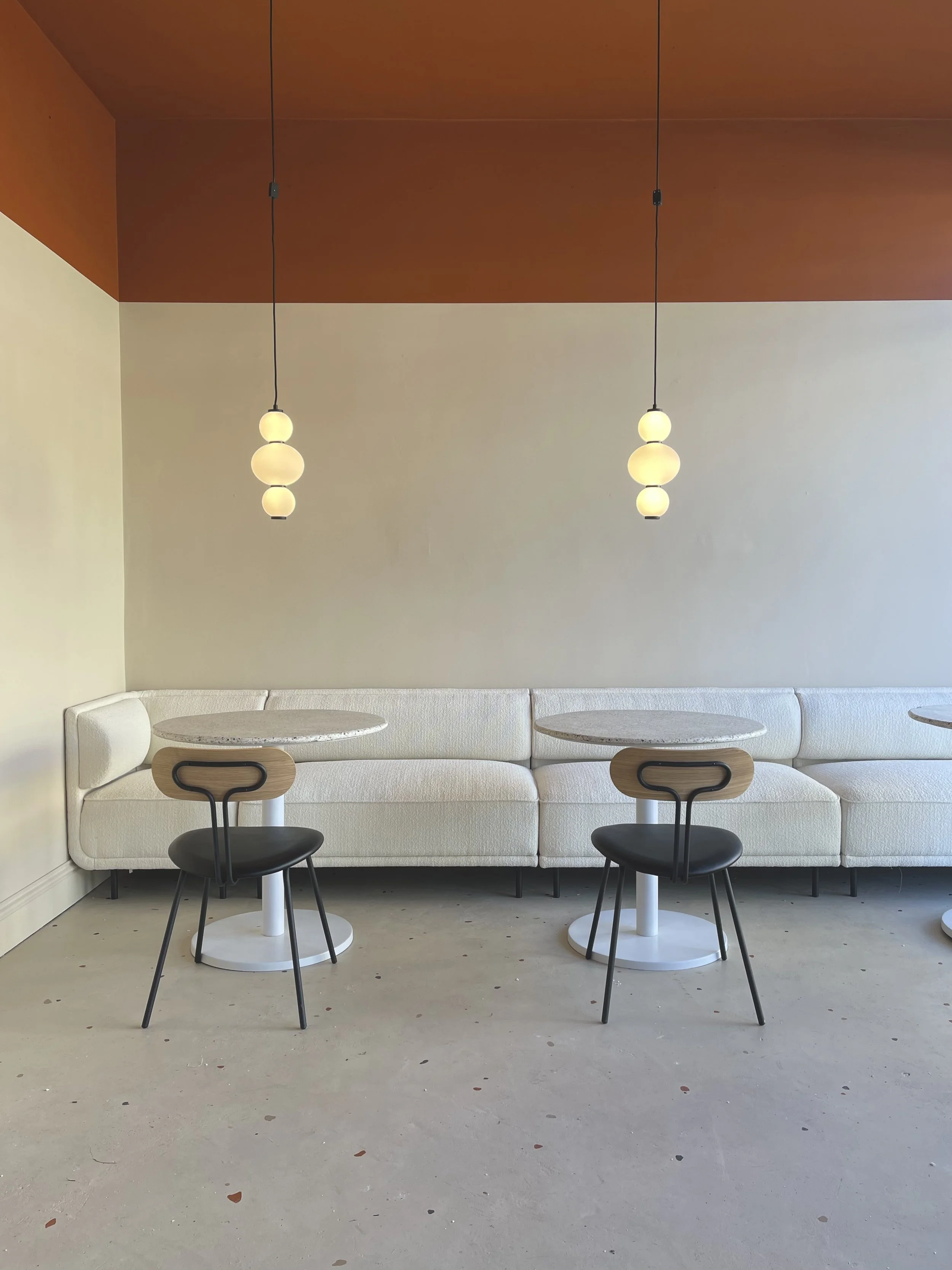 Modern interior with a long white cushioned bench against the wall, three hanging pendant lights above, and two small round tables with black chairs in front. The wall has a two-tone design with beige on the lower part and reddish-brown on the upper part, and the floor is light gray with scattered small objects.