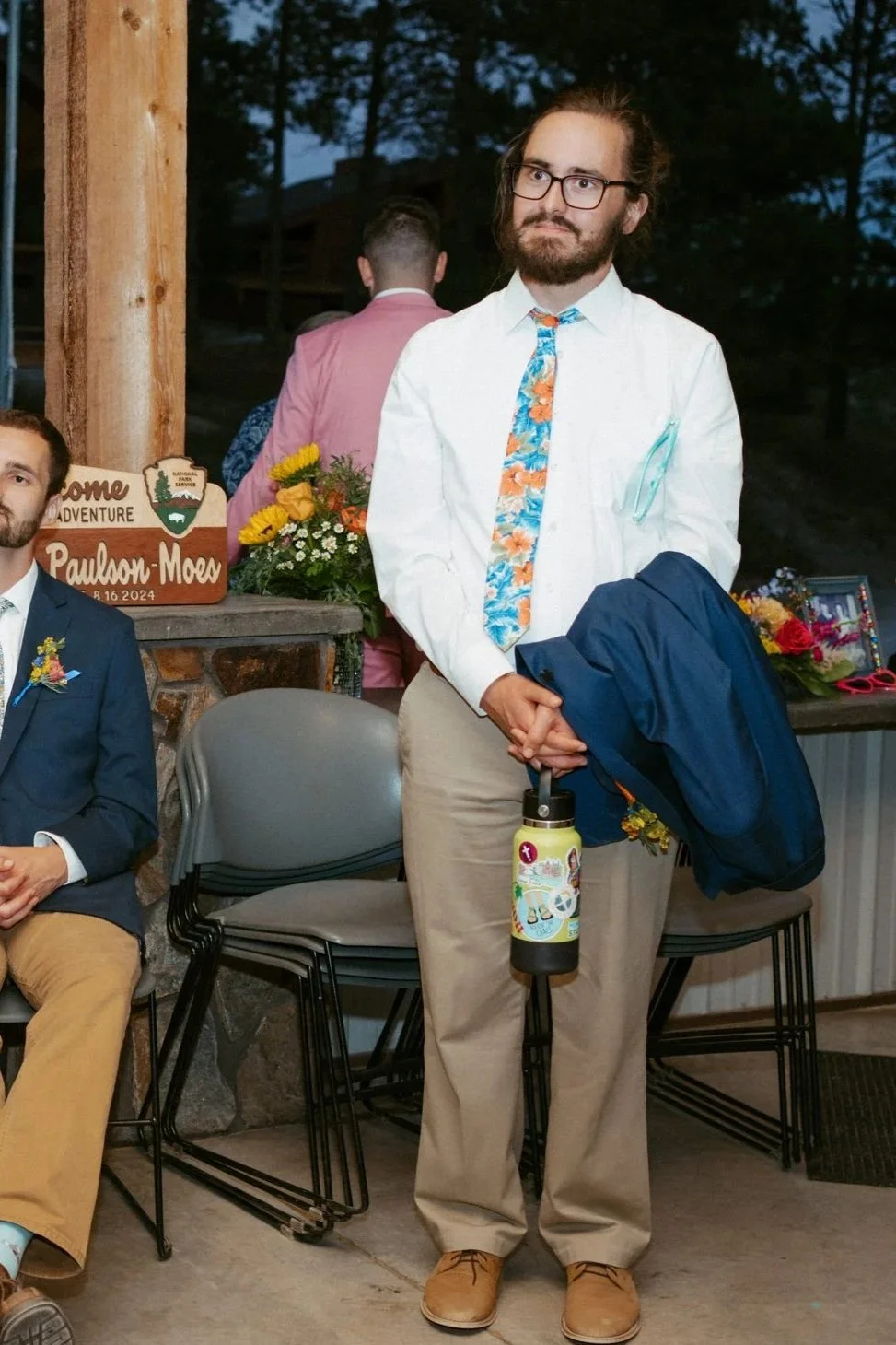 A man in beige pants, a white shirt, and a colorful floral tie holding a navy jacket and a water bottle, standing indoors during a social event.