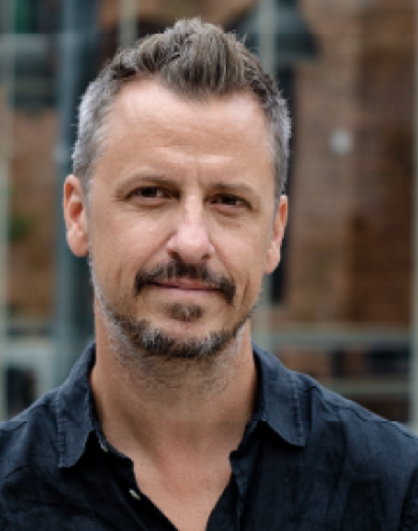 Close-up portrait of a middle-aged man with short, grayish hair and a beard, wearing a dark shirt outside in an urban setting with brick buildings and metal structures in the background.