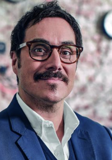 A man with dark hair, glasses, and facial hair wearing a blue blazer and white shirt, standing in front of a blurred pink and white background.
