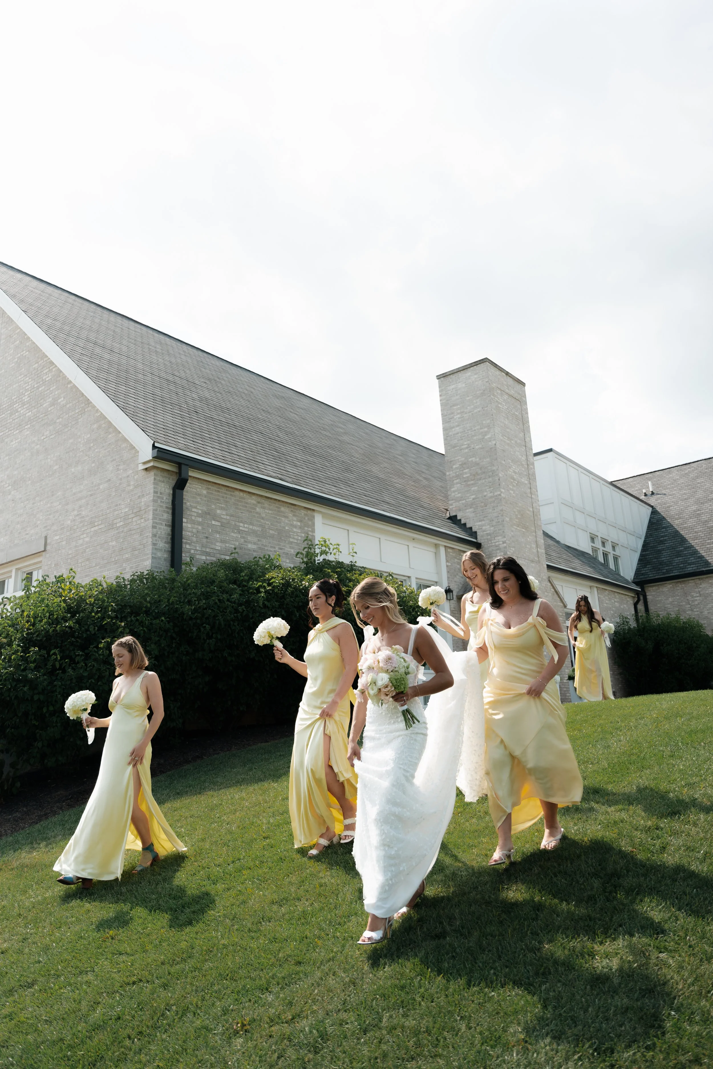 A bride in a white wedding dress and veil holding a bouquet of pink and white roses, walking on green grass with bridesmaids in light yellow dresses holding white flower bouquets behind her, near a brick building with a sloped roof and large windows.