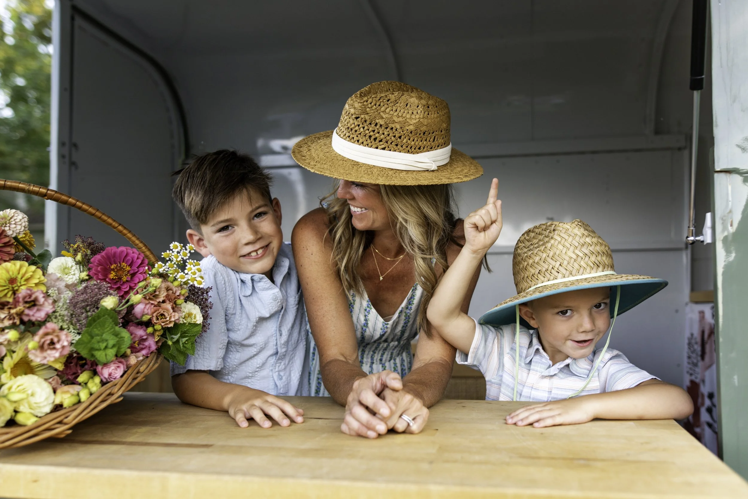 A woman with long blonde hair wearing a straw hat and two young boys, one wearing a straw hat with a blue ribbon and the other without, smiling and leaning on a wooden table inside a flower stand with a basket of colorful flowers nearby.