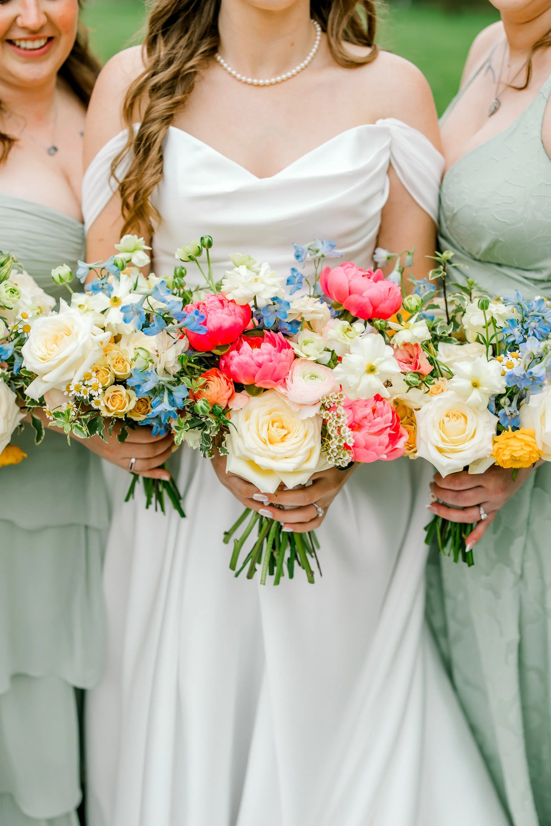 Close-up of a bride and two women holding a large bouquet of colorful flowers at a wedding, smiling and dressed in pastel dresses with necklaces.