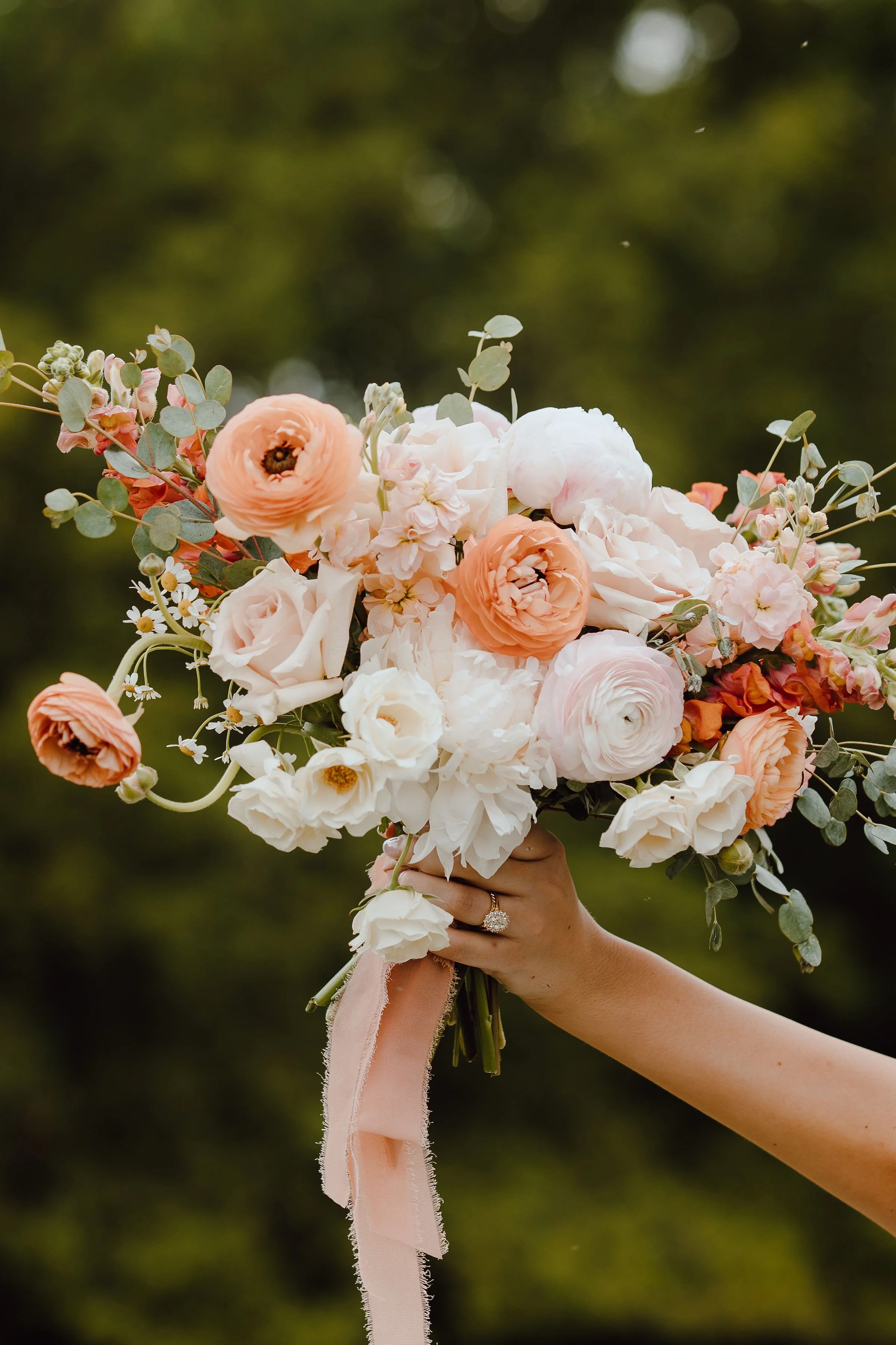 A person's hand holding a large bouquet of pink and white flowers with green foliage, tied with a pink ribbon, against a blurred green outdoor background.