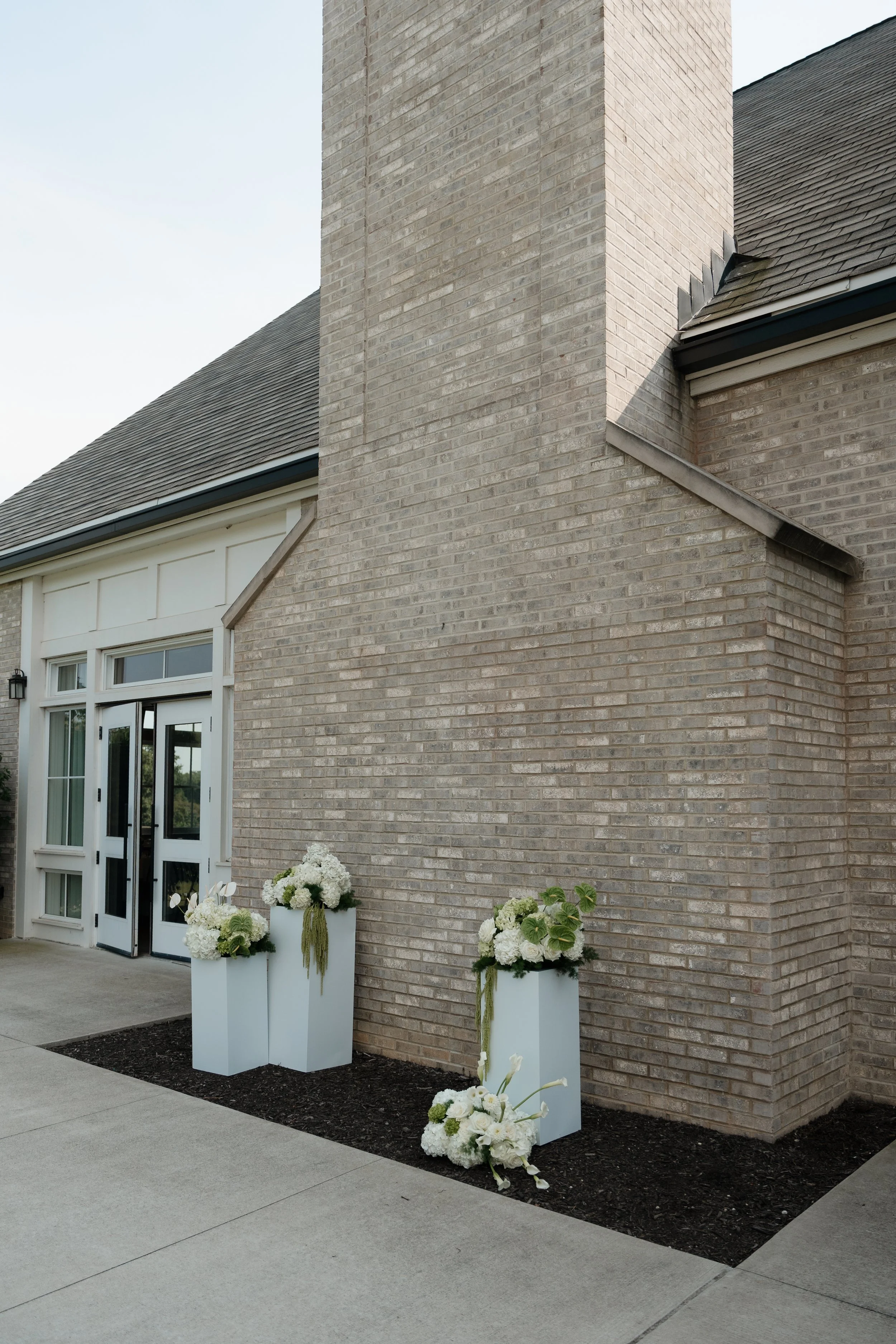 Exterior of a modern brick house with white flowers in tall white planters near the entrance, and some flowers on the ground.