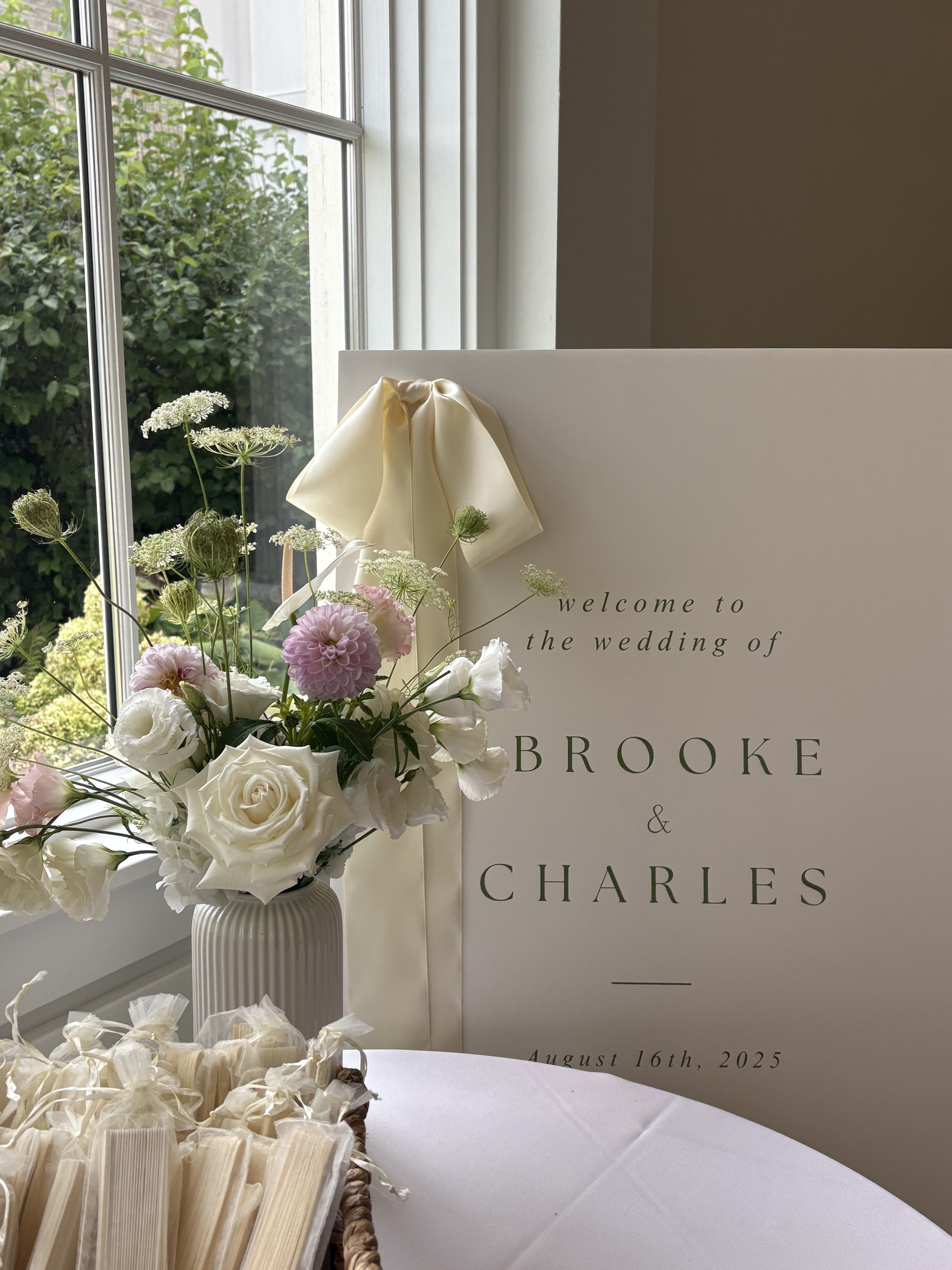 A wedding welcome sign with floral arrangements, set near a window with greenery outside. The sign reads "welcome to the wedding of Brooke & Charles" and has the date August 16th, 2025.