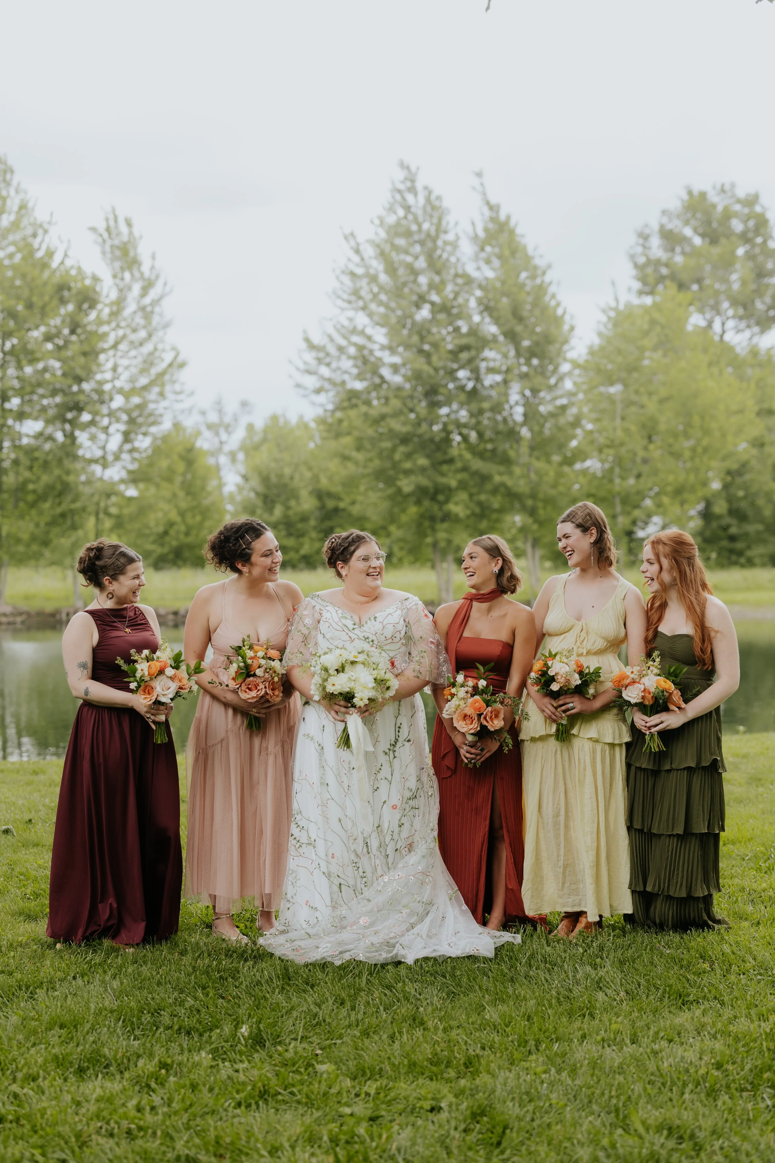 A group of seven women dressed in colorful dresses standing outdoors near a lake, holding bouquets of flowers, smiling, and looking at each other, with trees and a cloudy sky in the background.