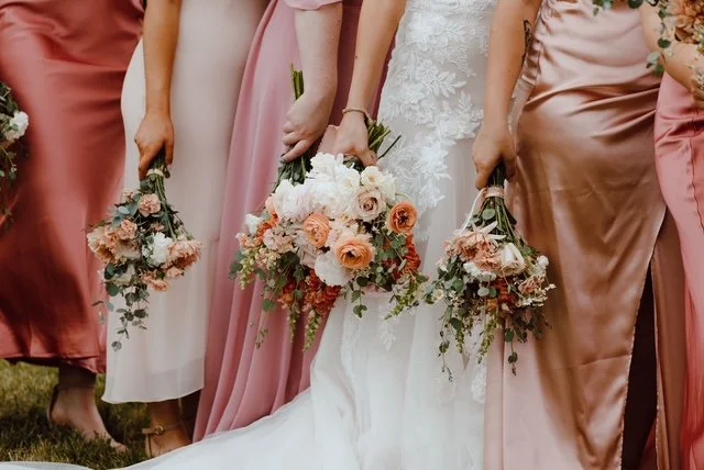 Close-up of bridesmaids in pink and white dresses holding bouquets of mixed flowers including roses, positioned outdoors.