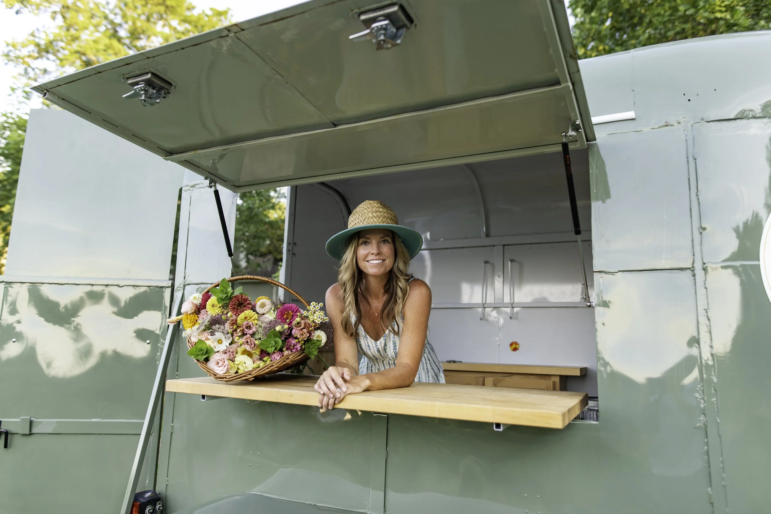 A woman with long hair, wearing a striped summer dress and a wide-brimmed hat, is standing behind a wooden counter inside a vintage green trailer, holding a large basket of colorful flowers, smiling at the camera, with trees in the background.