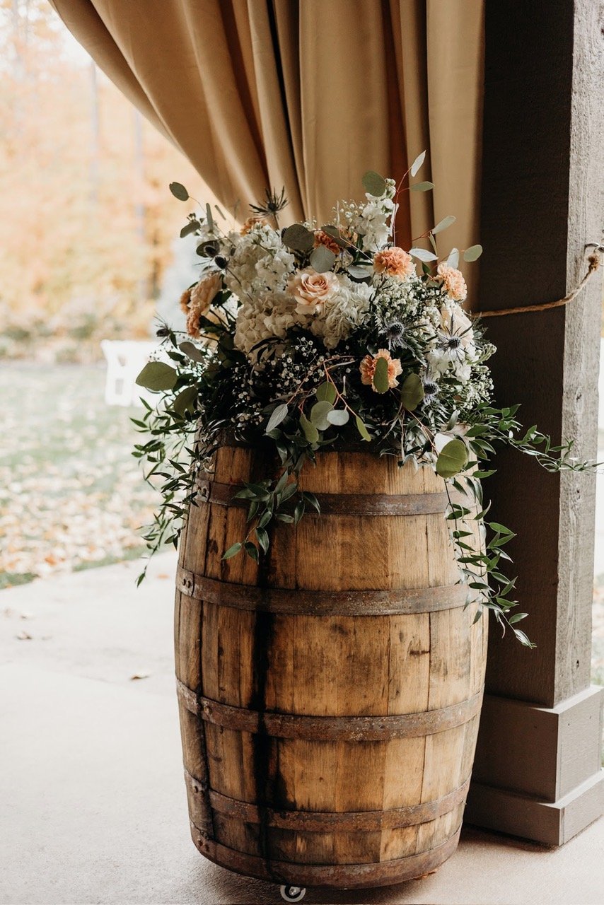 A rustic wooden barrel used as a stand for a bouquet of white and peach flowers and green foliage, placed beside a view outside with autumn trees and a gold curtain.