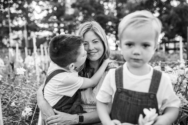 A woman with two young boys outdoors in a garden, one boy kissing her cheek and the other standing nearby, blurred in the foreground.