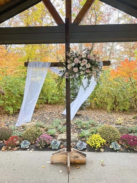 Outdoor wedding arch decorated with white sheer fabric and a floral arrangement, set against autumn foliage.