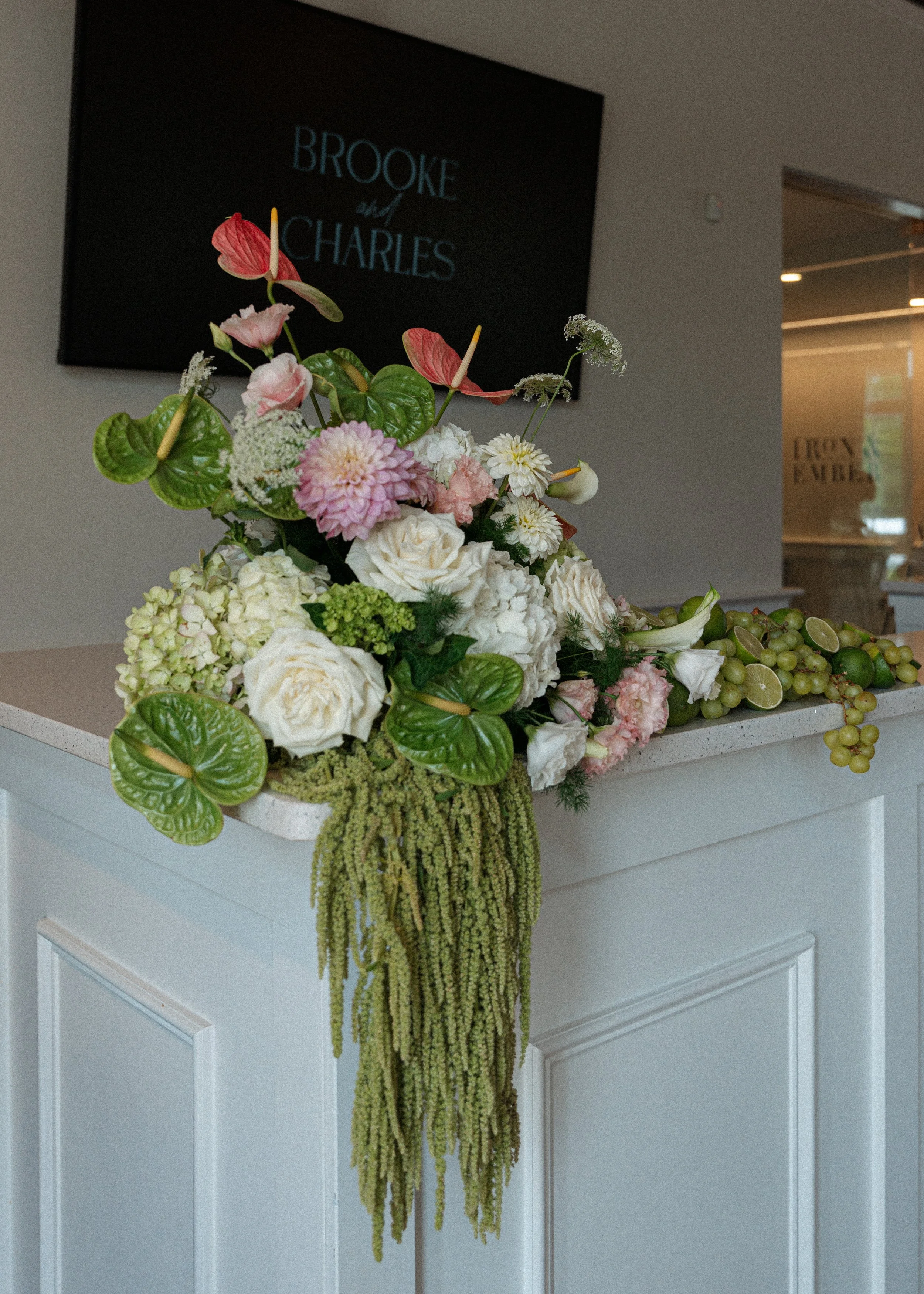 Elegant floral arrangement with pink, white, and green flowers, accented with green hanging amaranthus, placed on a white dresser in front of a wall with artwork and a sign.