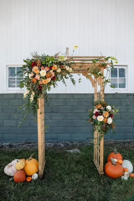 Decorative wooden arch with floral arrangements on top and sides, surrounded by pumpkins on the ground.
